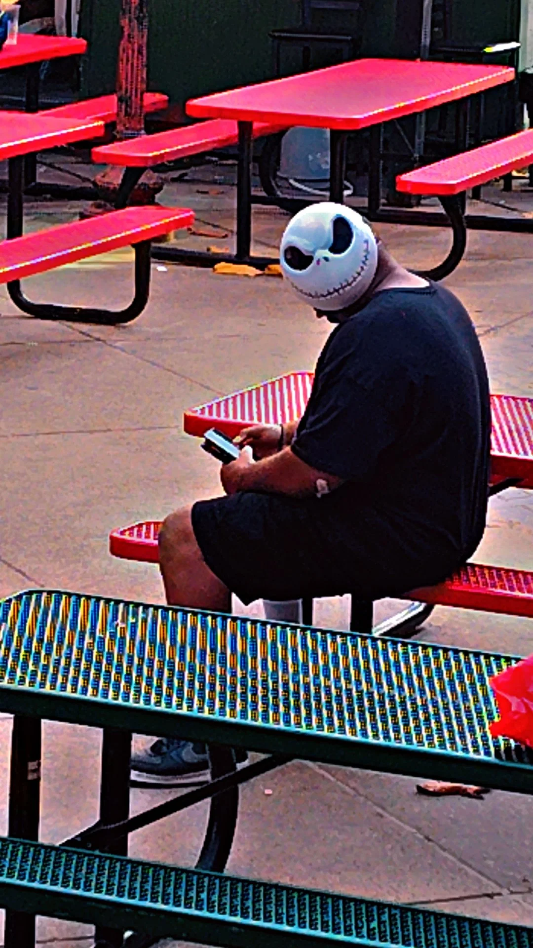 Person wearing a Jack Skellington mask sitting on a red outdoor bench, looking at their phone, surrounded by red and blue picnic tables.