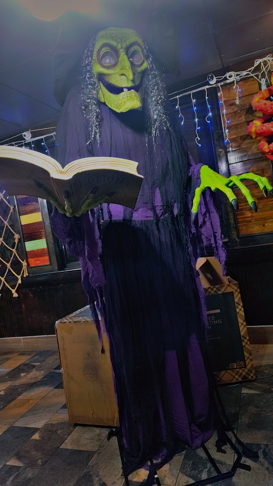 A life-sized Halloween decoration of a witch with a green face and long, dripping hair, wearing a purple dress, holding an open book, and standing on a platform in a decorated room.