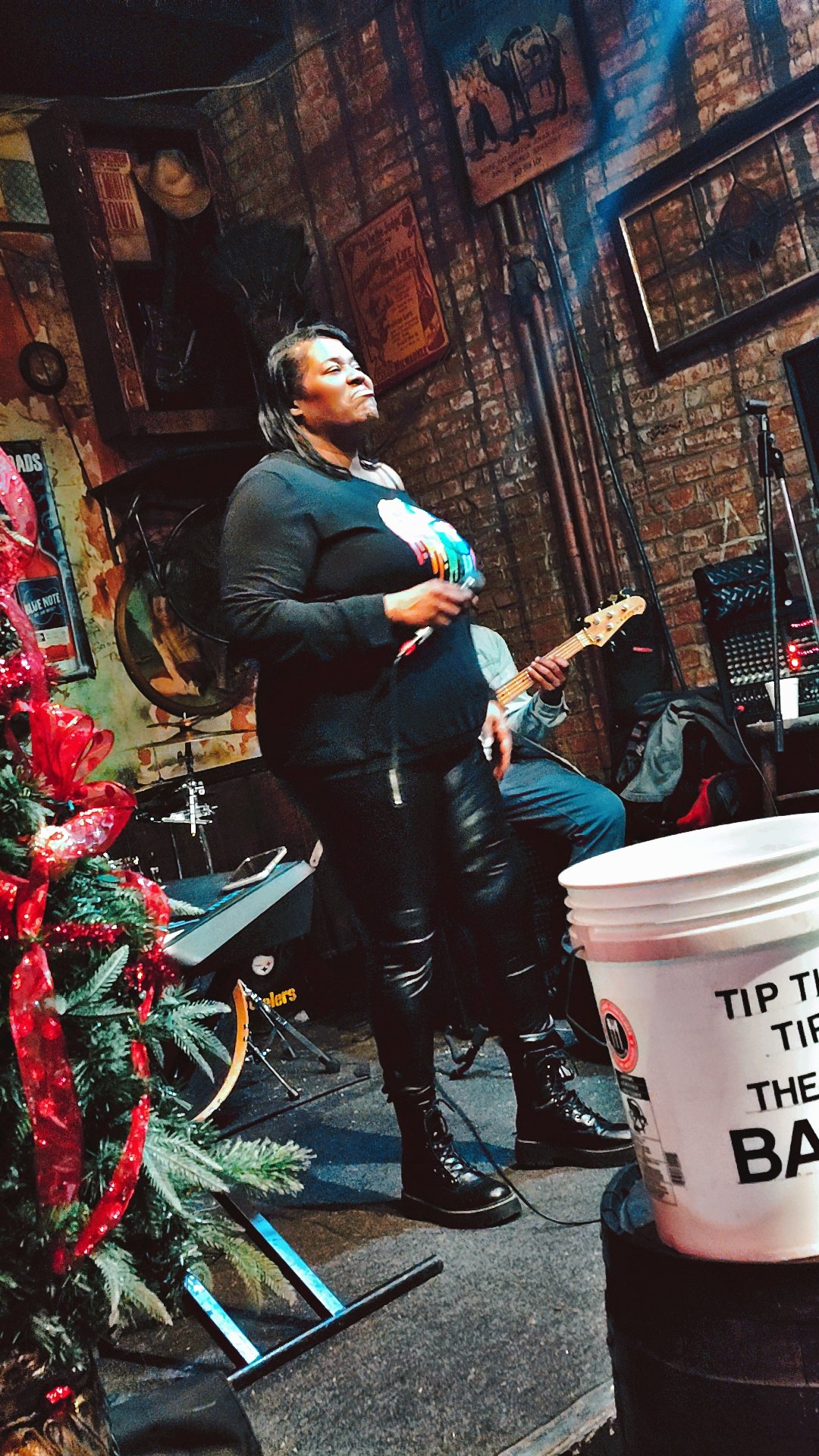A woman singing into a microphone while holding a guitar at a bar or music venue, with a Christmas tree decorated with red bows in the foreground and brick walls with posters and decorations.