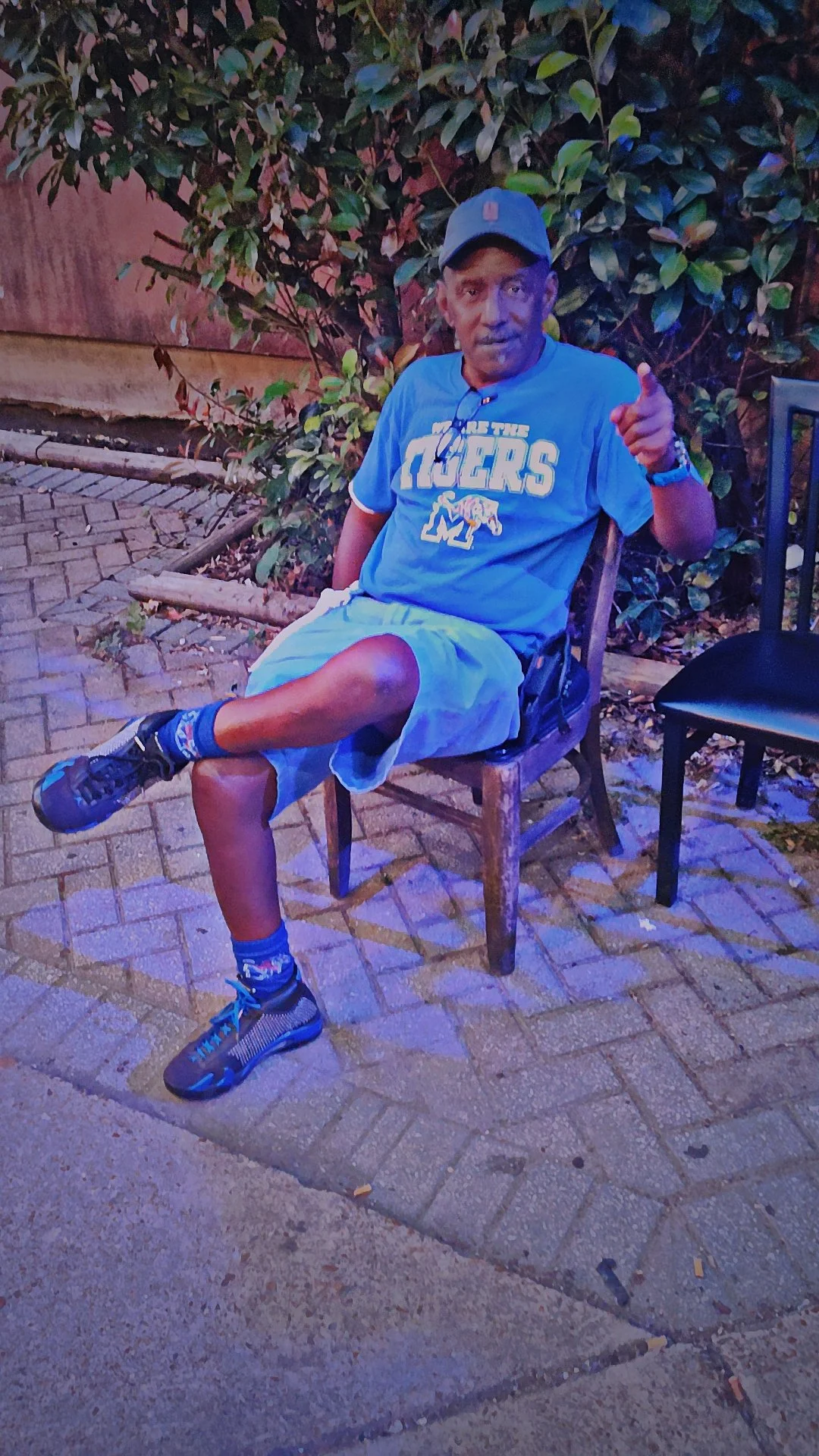 A man sitting on a wooden chair outdoors at night, pointing towards the camera, wearing a blue sports t-shirt, gray shorts, a cap, and sneakers, with a lush green bush and a fence in the background.