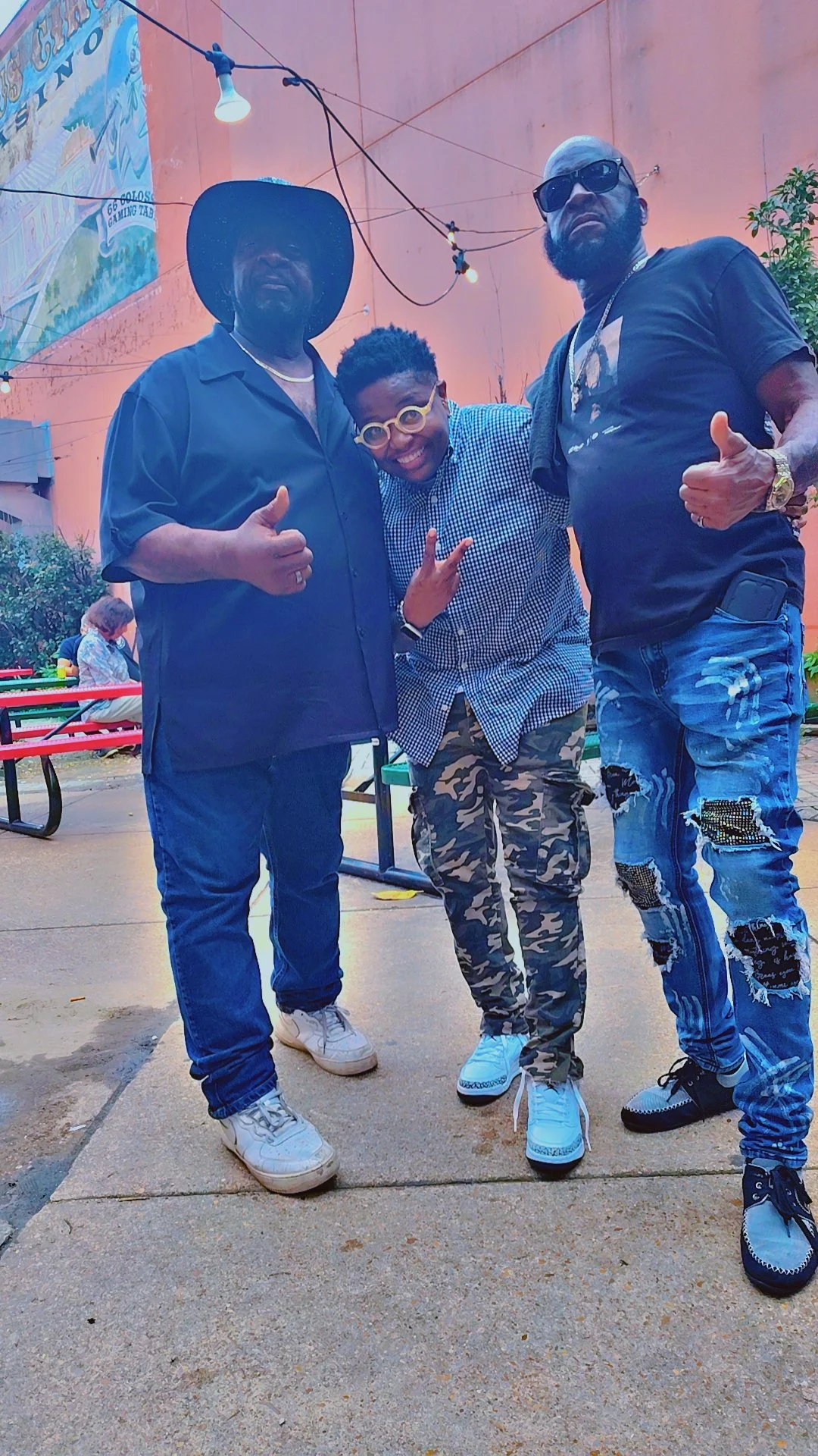 Three people standing outdoors on a sidewalk, posing for a photo. The person on the left is wearing a wide-brimmed hat, dark shirt, and jeans. The person in the center is dressed in a blue checked shirt and camouflage pants, making a peace sign. The 