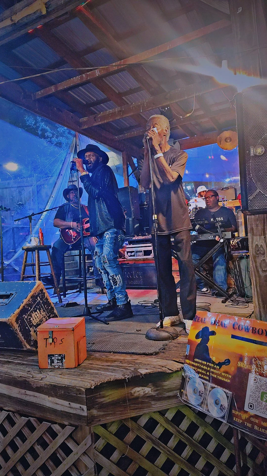 Musicians performing on a wooden stage with a man singing into a microphone, a woman playing guitar, and other musicians in the background. There is a promotional sign at the front of the stage with CDs and text promoting a concert event.