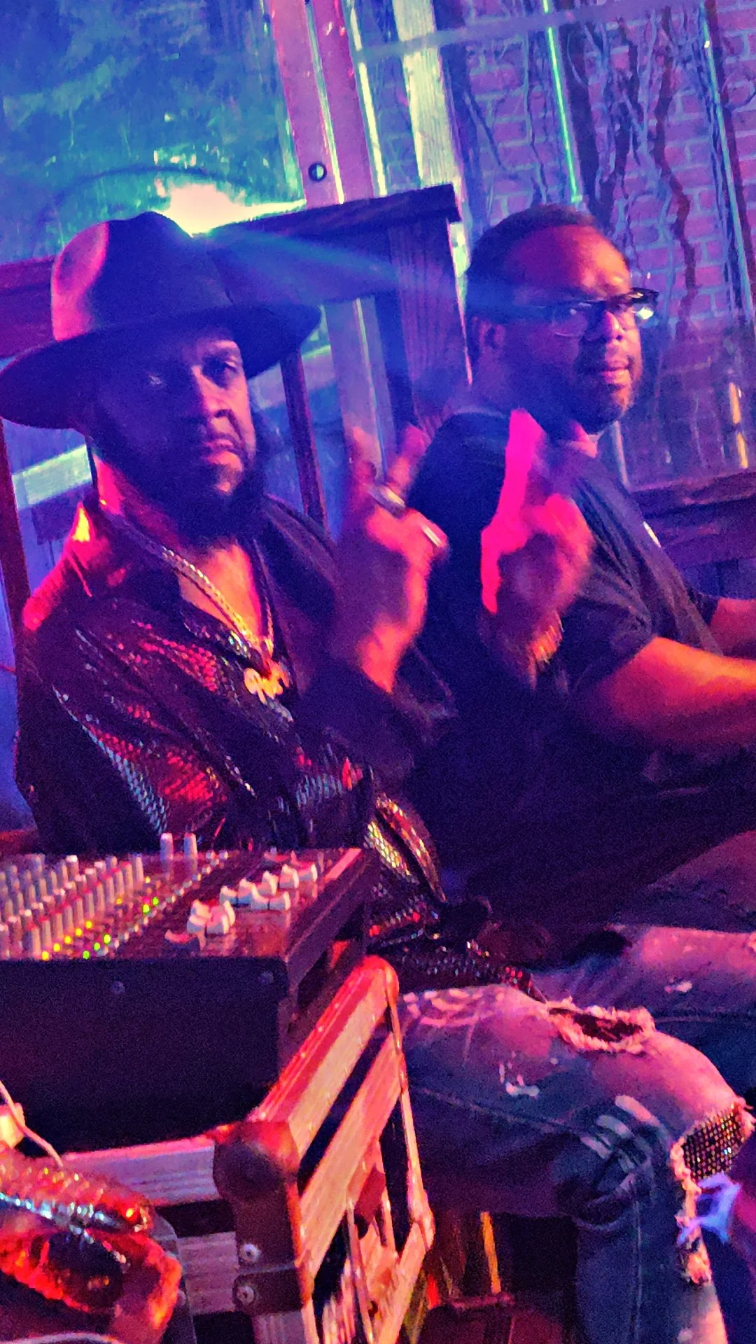 Two men sitting in a vibrant, colorful nightclub or bar setting. One man is wearing a large hat and jewelry, and the other is wearing glasses. There is DJ equipment in the foreground.