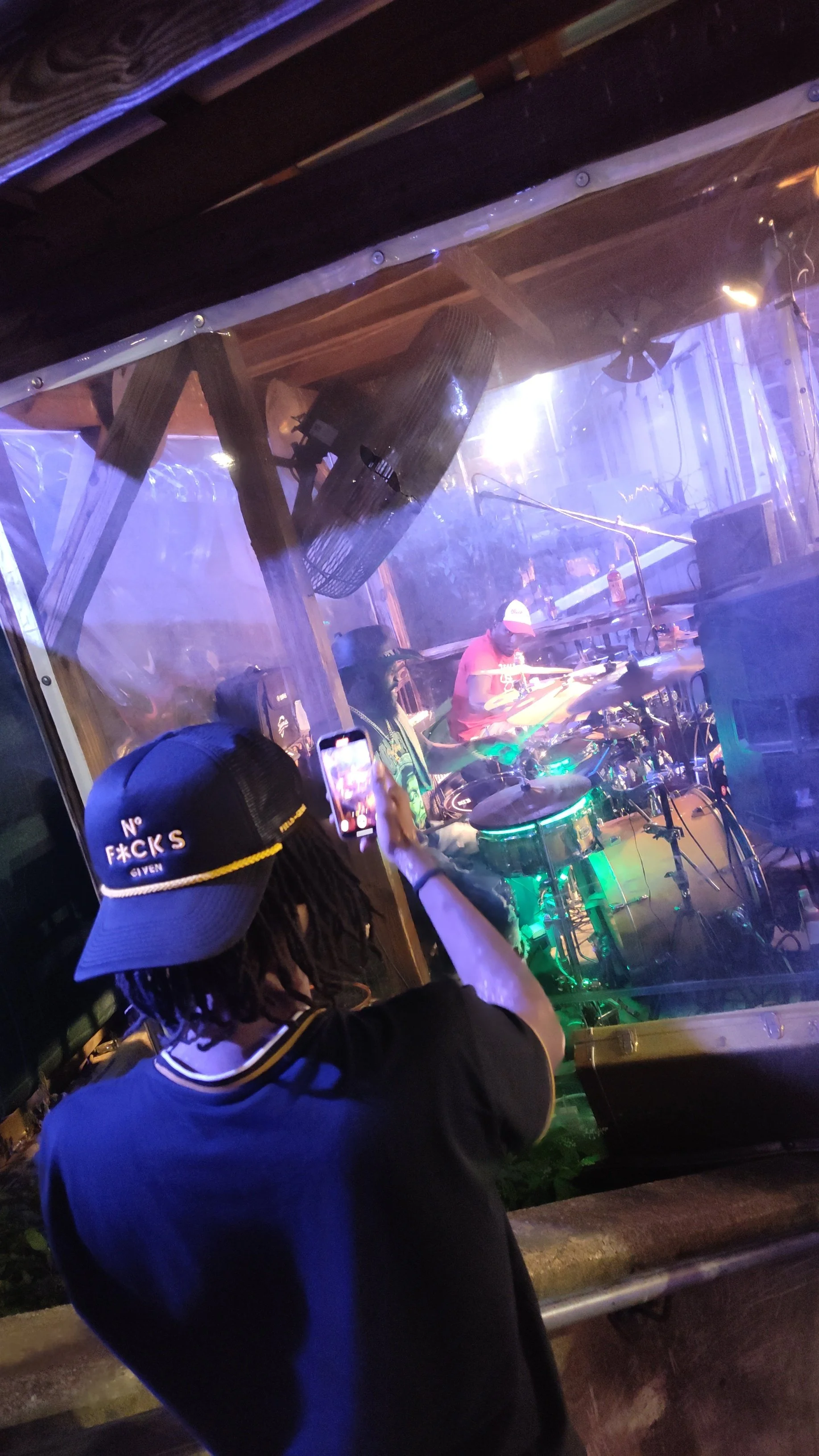 Person taking a photo of a drummer playing in a live music venue, with colorful lighting and a plastic window barrier.
