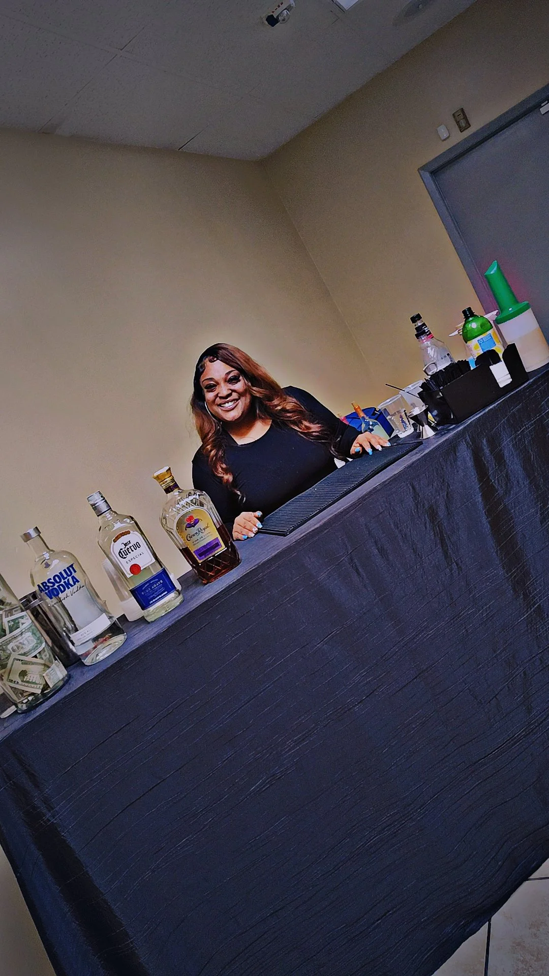 A woman behind a bar with liquor bottles and cash on the counter, smiling at the camera.