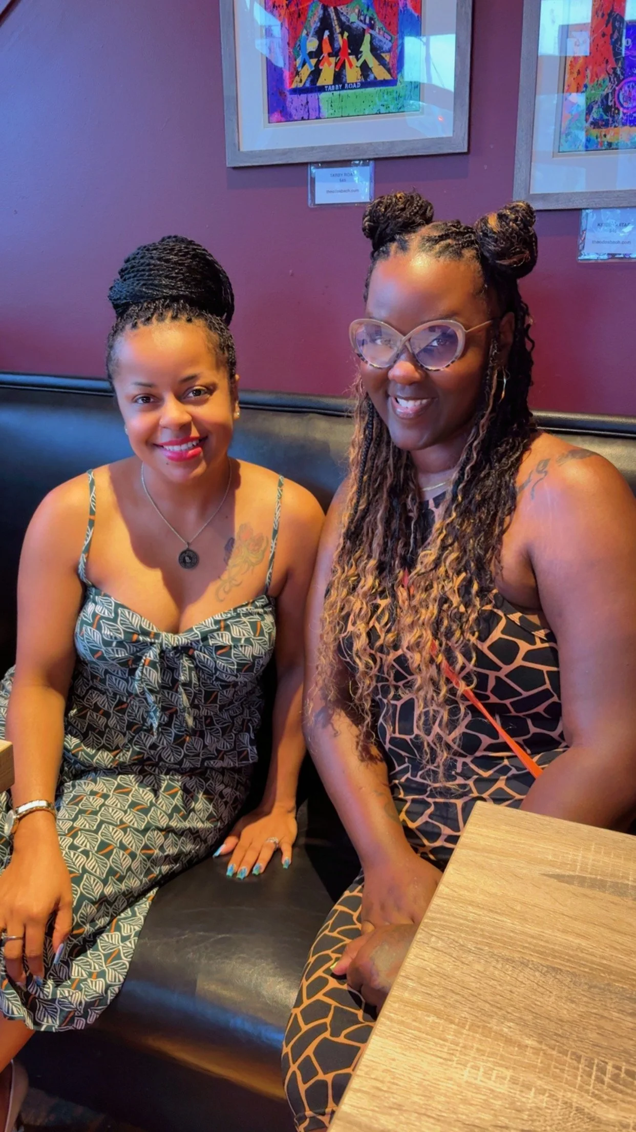 Two women sitting at a restaurant table, smiling at the camera. The woman on the left has dark hair styled in braids, wearing a sleeveless patterned dress, and jewelry. The woman on the right has long, curly hair styled in buns with glasses and a sle