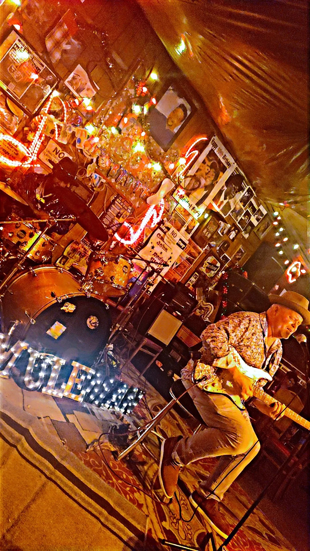 A musician playing an electric guitar on a stage decorated with Christmas lights, photos, and posters in a cozy, cluttered bar or club.