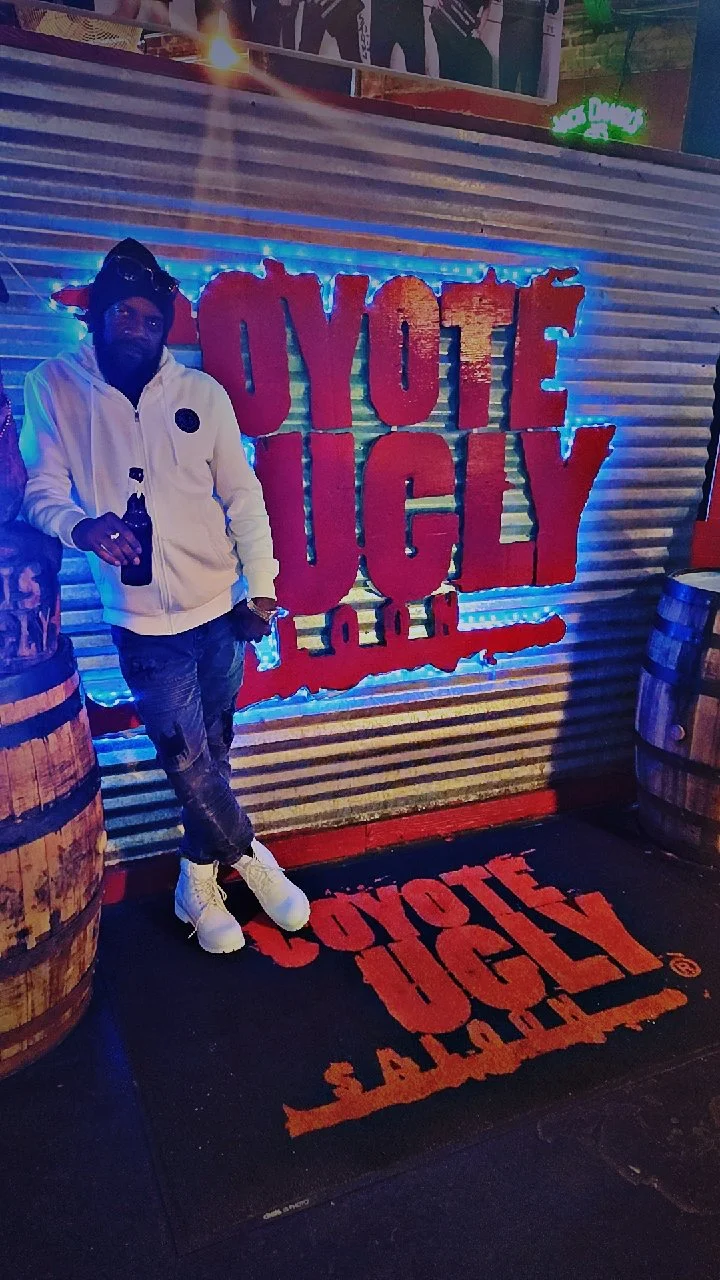 Person standing in front of a sign that reads 'OYO TE JUGY' with neon lights, surrounded by barrels.