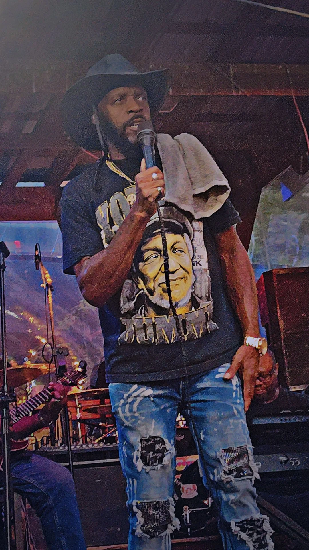 A man wearing a wide-brimmed hat, a black T-shirt with a graphic of an Indigenous person, and ripped jeans, is holding a microphone and standing on a stage with musical instruments in the background.