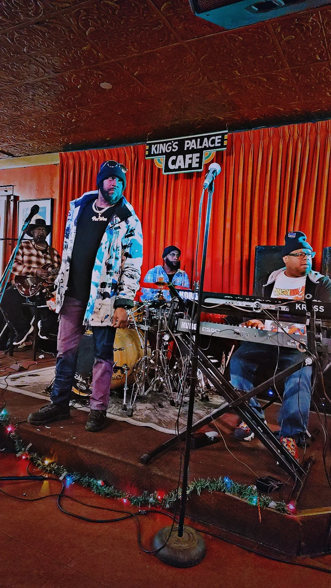 A band performing on stage at King's Palace Cafe, with three musicians playing instruments and one person singing or speaking into a microphone, decorated with red curtains and holiday lights.