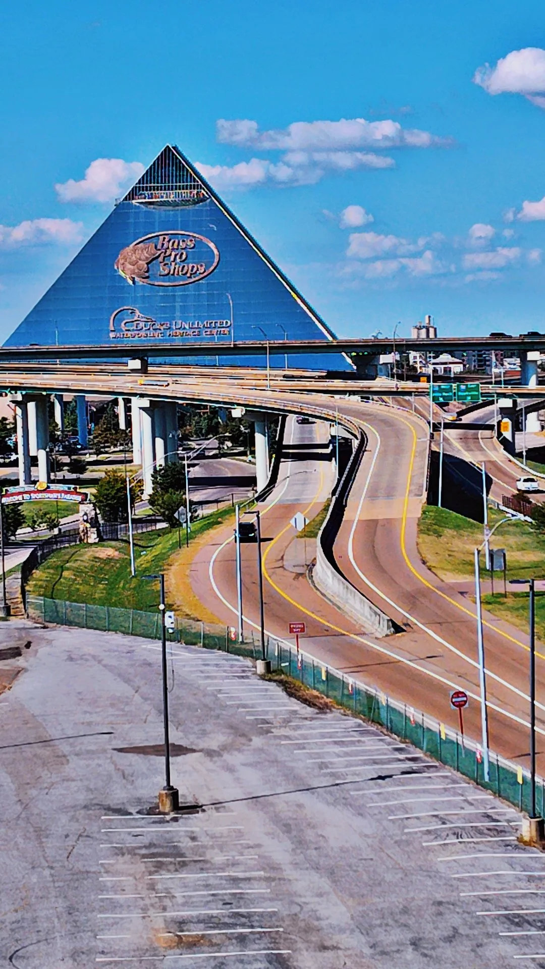 Empty parking lot with a road curving under a highway overpass, leading to a large, pyramid-shaped building with a Bass Pro Shops sign on it in the background under a blue sky with some clouds.