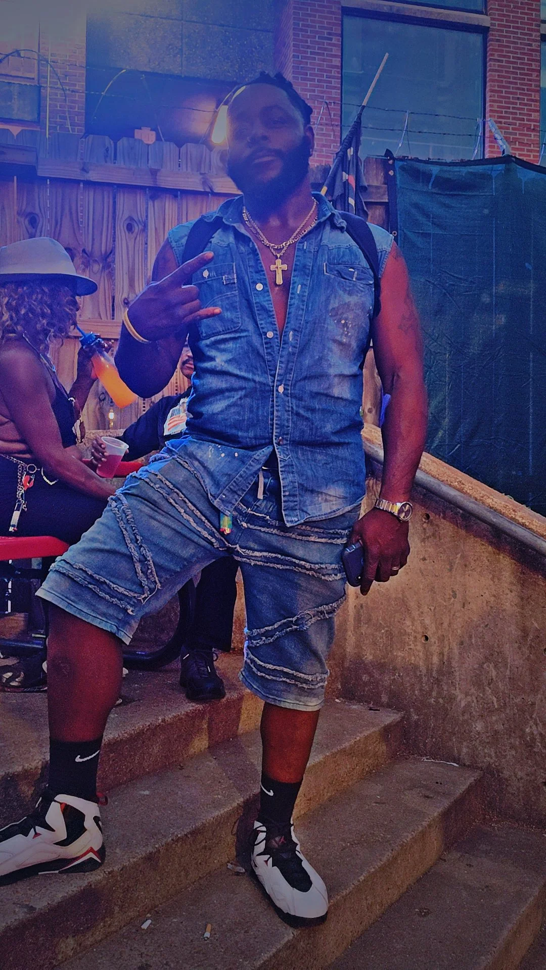 A man posing with a gesture, wearing a denim vest, denim shorts, sneakers, and jewelry in an outdoor setting at night.