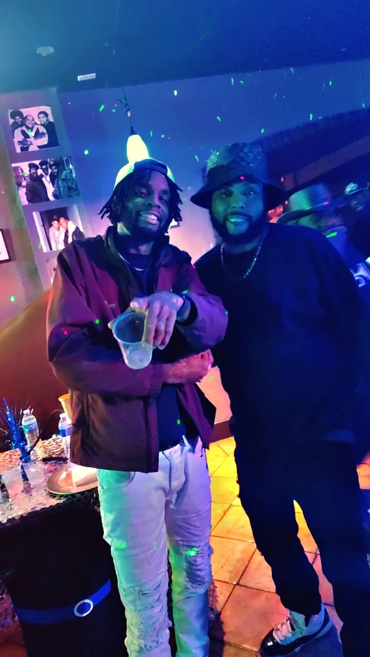 Two men are posing and smiling at a party or nightclub with colorful lights, one holding a drink, the other wearing a bucket hat and chain, with a photo collage on the wall behind them.