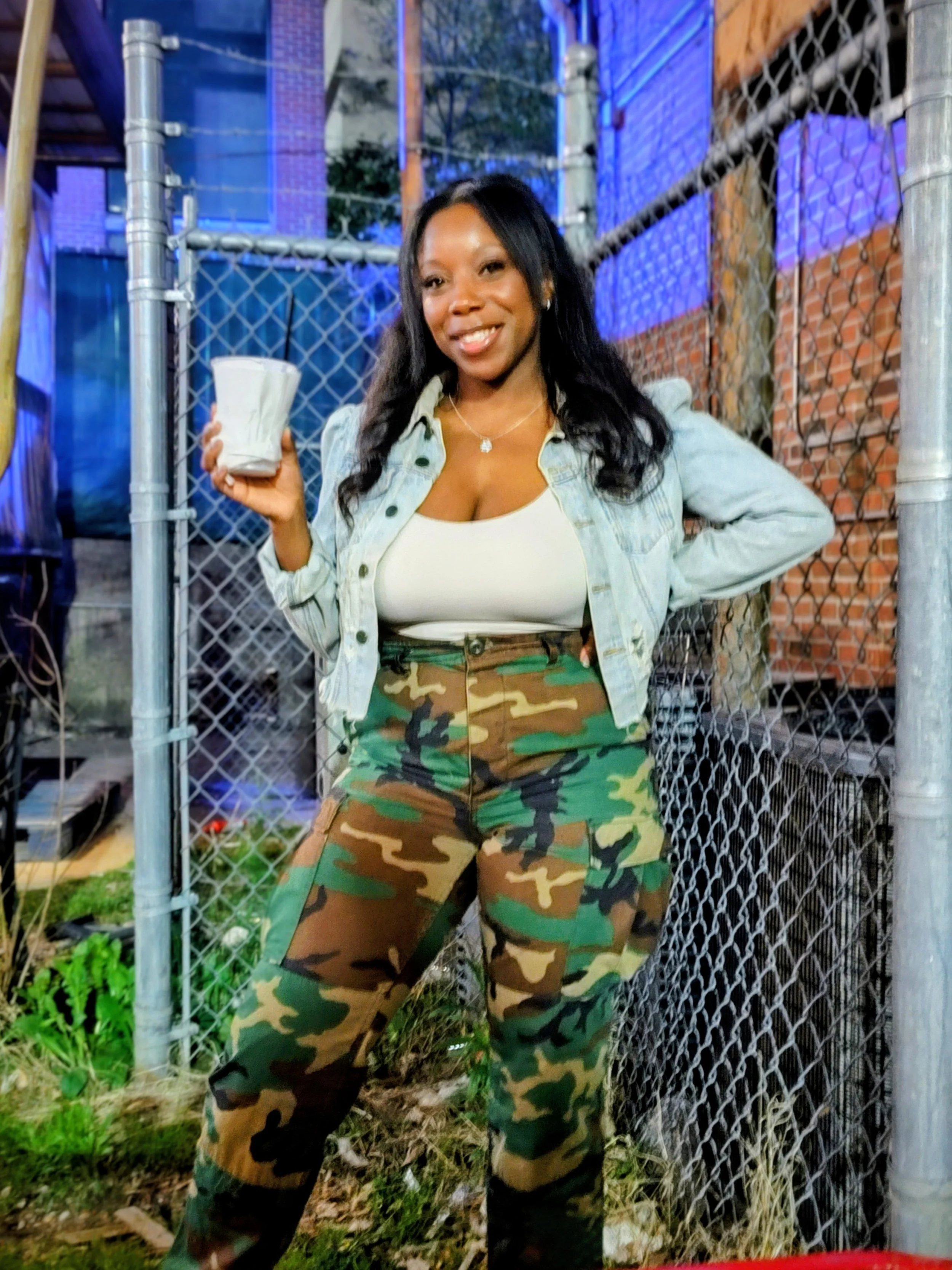 A woman smiling and holding a white cup stands outdoors near a chain-link fence, wearing a light denim jacket, a white top, and camouflage pants, with a background of a brick building and greenery.