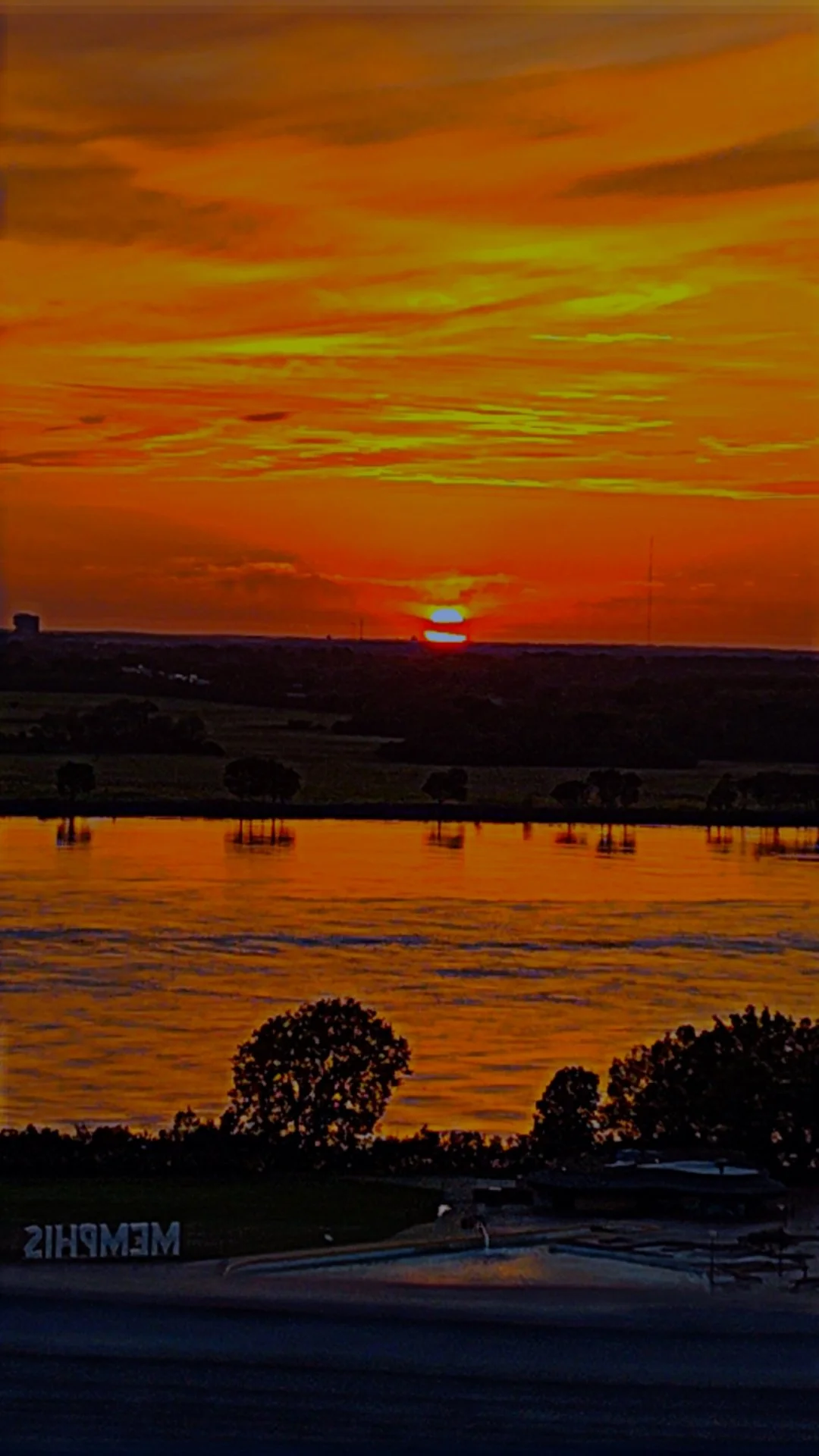 A vibrant sunset over a body of water, with a bright orange and yellow sky, silhouetted trees, and boats reflected in the water.