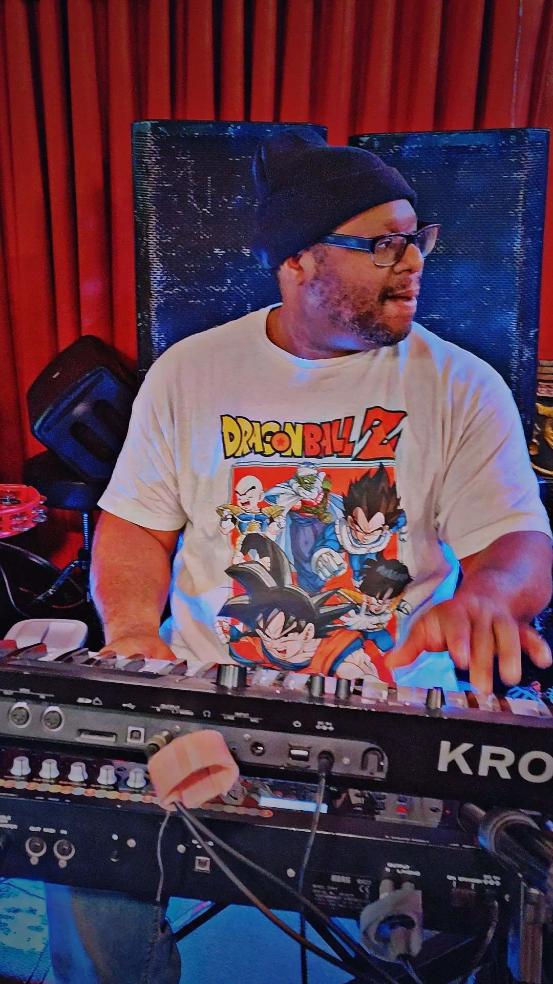 A man wearing a black beanie, glasses, and a Dragon Ball Z T-shirt playing a keyboard, seated in front of a red wooden wall.