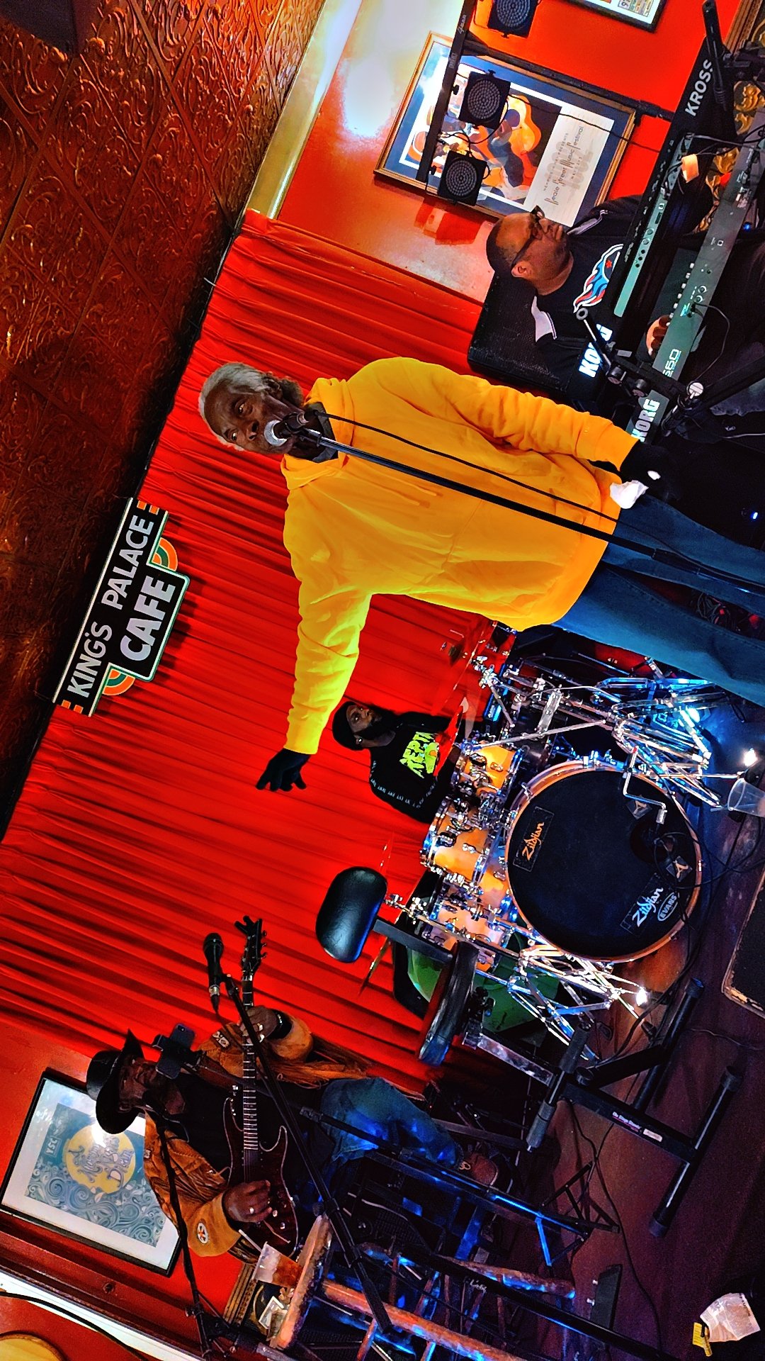 A musical performance on a small stage with a red curtain backdrop at King’s Palace Cafe. A man in a yellow hoodie is singing into a microphone, while a drummer and a guitarist are playing nearby. The setting appears cozy and lively.