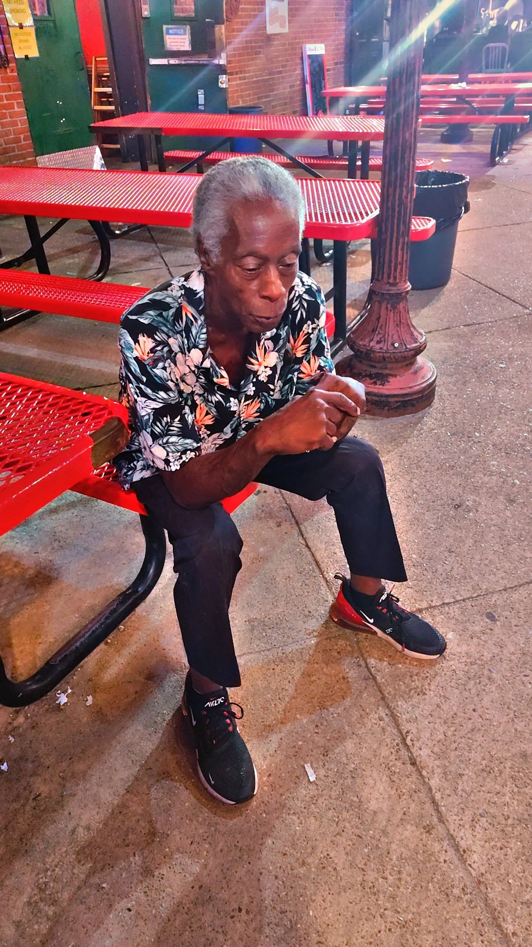 An elderly man with gray hair sitting alone on a red metal bench outside a restaurant or bar at night. He is wearing a black and white floral shirt, black pants, and black and red athletic shoes. The setting is urban with brick walls, a lamppost, and