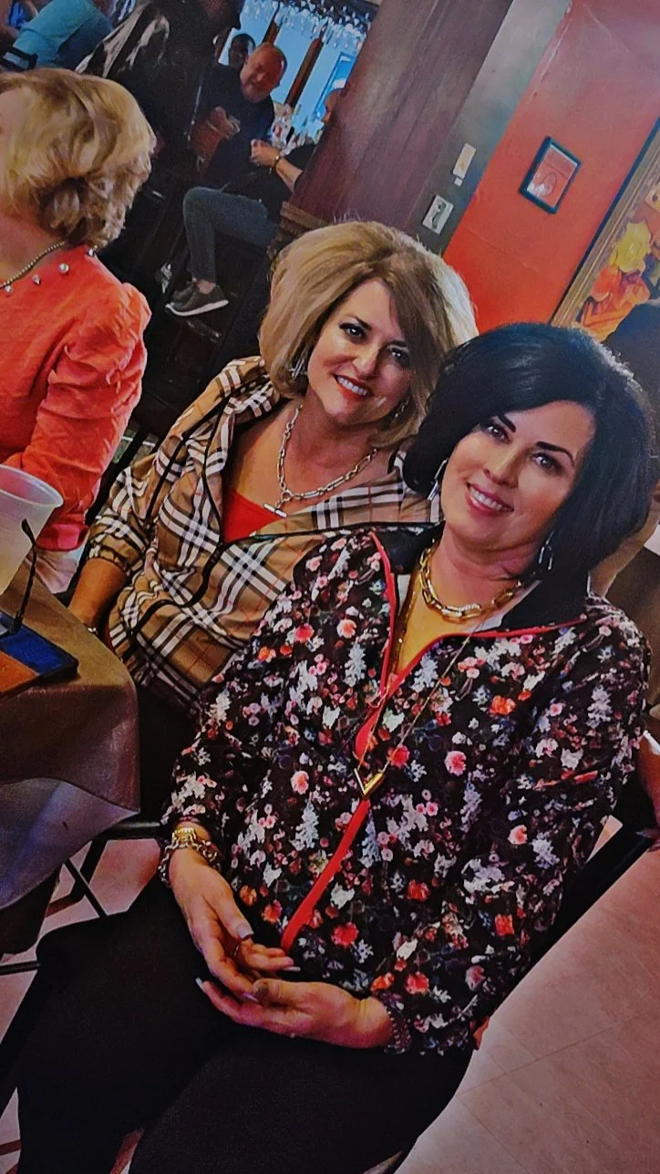 Two women smiling and sitting at a restaurant or bar, with other people in the background, one woman wearing a floral jacket and the other wearing a plaid shirt.