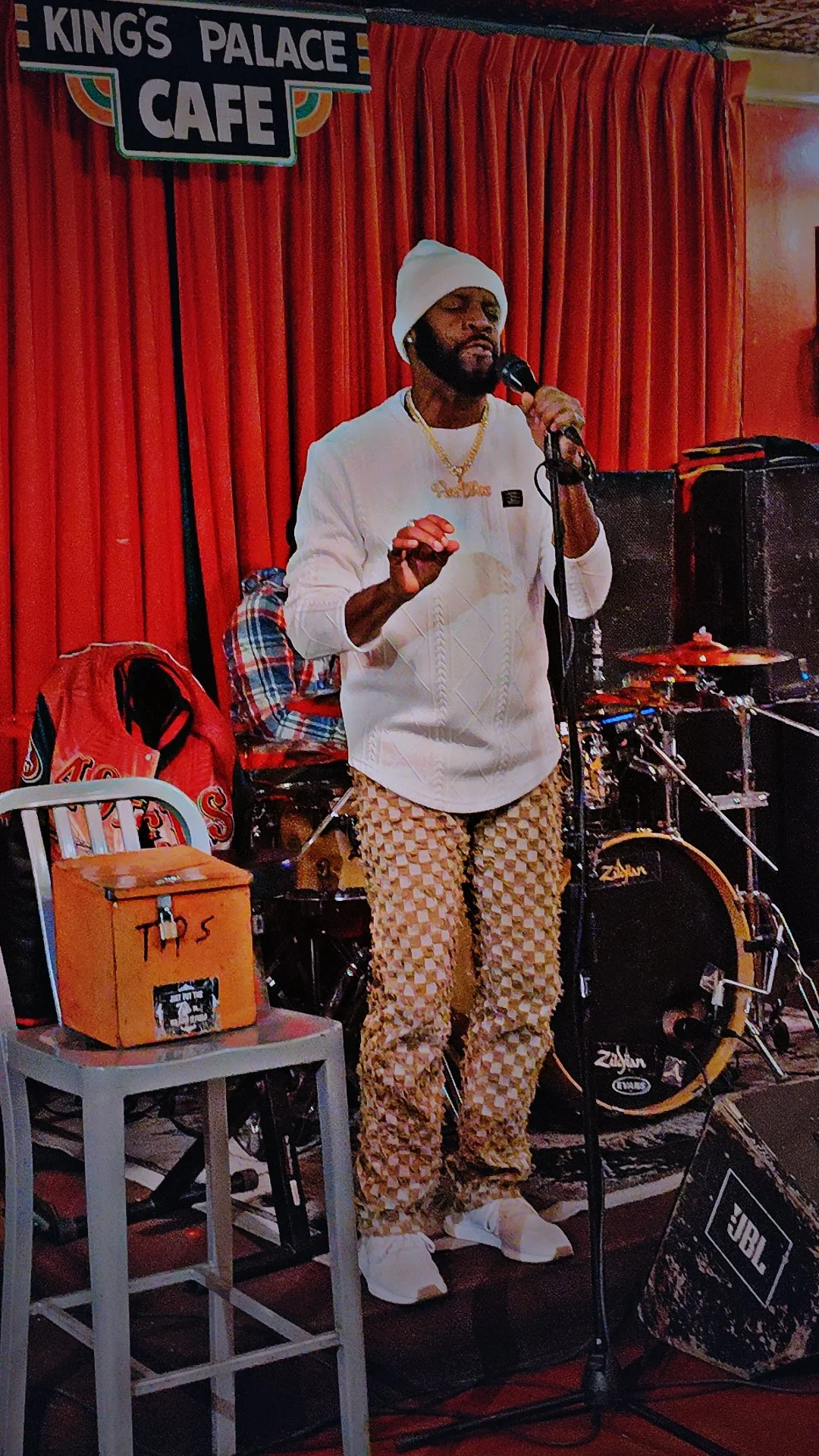 A man in patterned pants, white sweater, and white beanie singing into a microphone on a stage with red curtains, a drum set, and sound equipment.