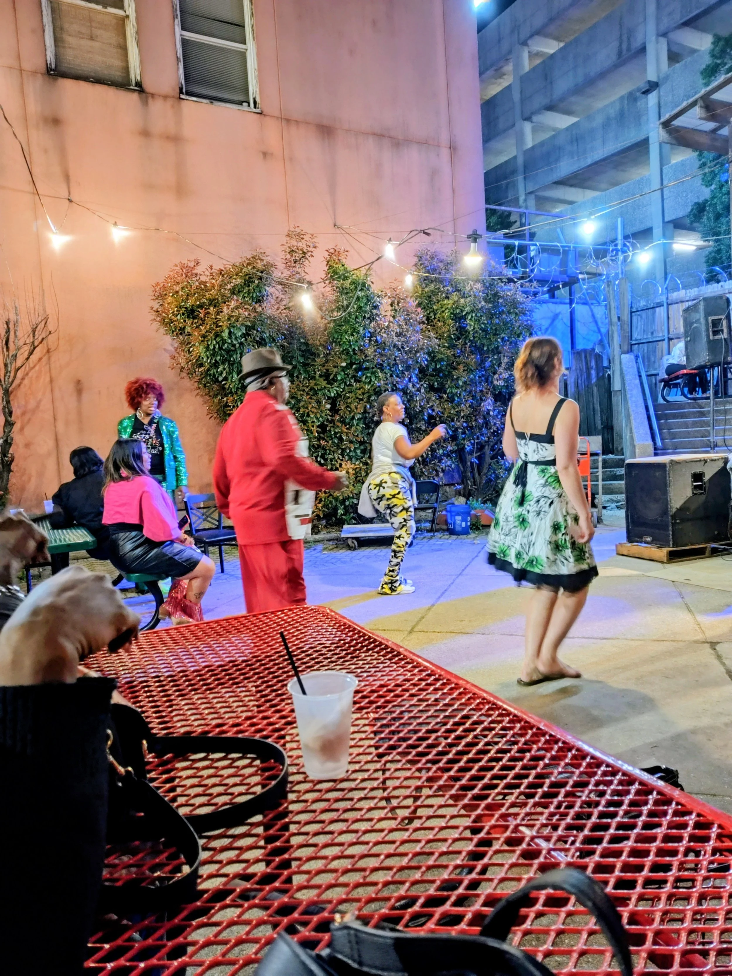 People dancing in an outdoor patio space with string lights, a pink wall, and a large bush. There are several chairs with some people seated, and a dog with a drink on a red metal table in the foreground.