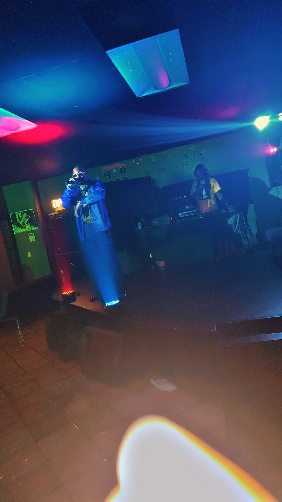 Two performers on a stage in a dimly lit nightclub, one with a microphone and the other operating DJ equipment, illuminated by colorful laser lights.