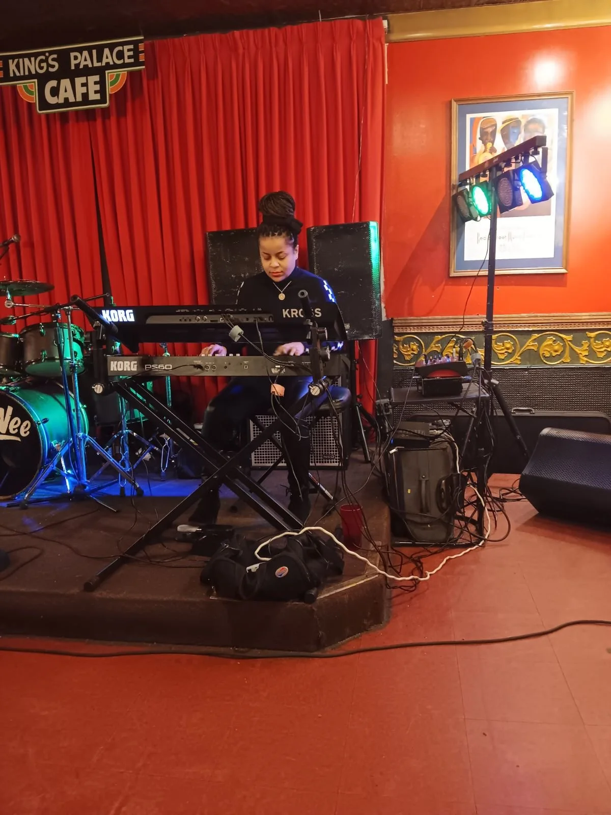 A woman playing a keyboard on stage at a live music venue, with drums and stage lighting, red curtains, and a framed picture on the wall behind her.