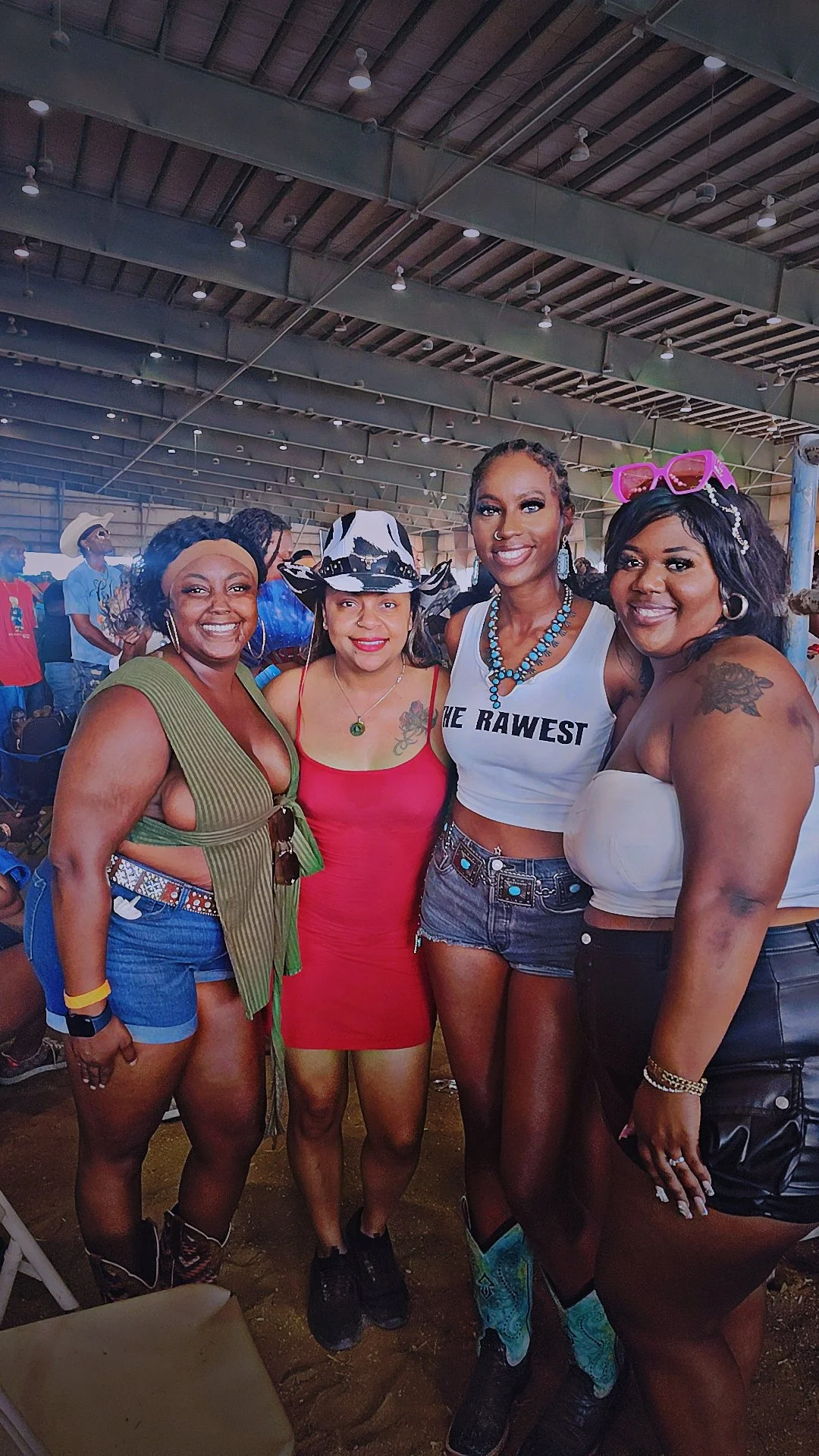 Four women smiling and standing together inside a building with a metal roof, dressed in casual or festival attire.