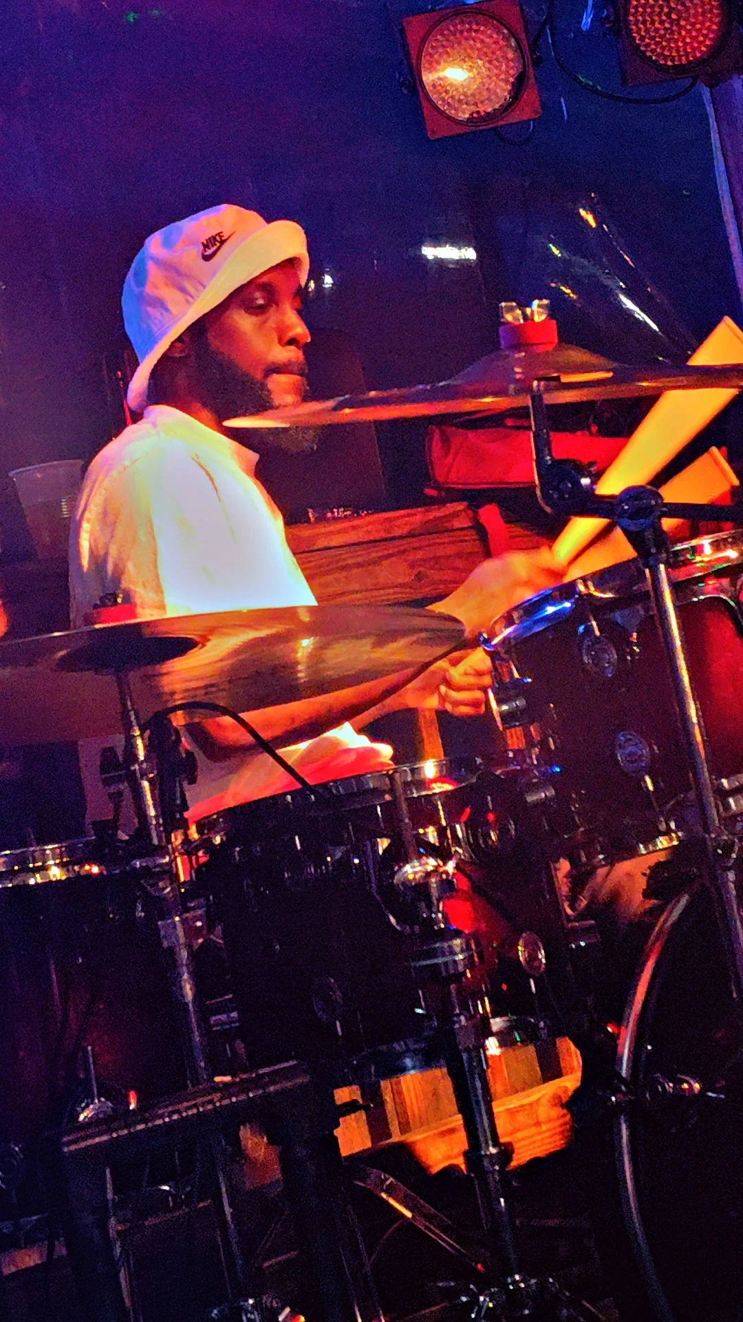 A man playing a drum set on stage, wearing a white Nike bucket hat and white shirt, with lighting creating a vibrant atmosphere.