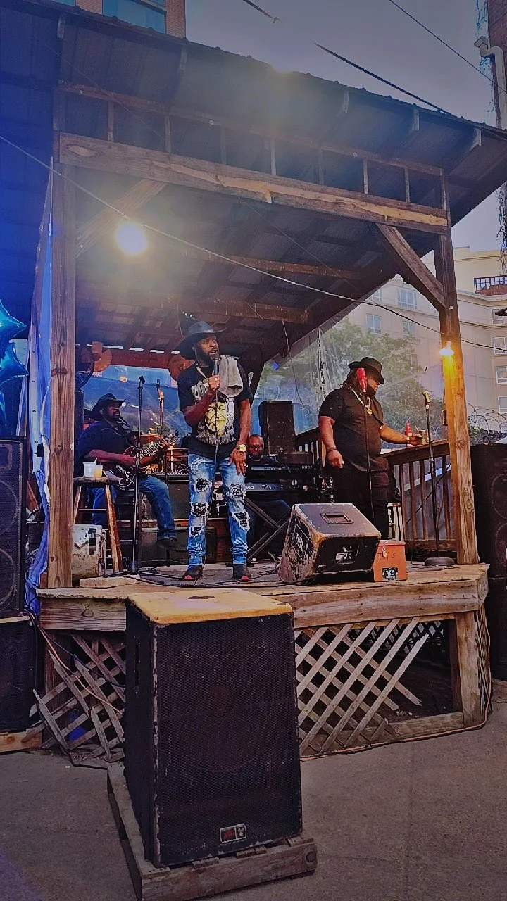 A live music performance on a small wooden outdoor stage with four musicians, one singing, two playing guitar and keyboard, in an urban setting during dusk.