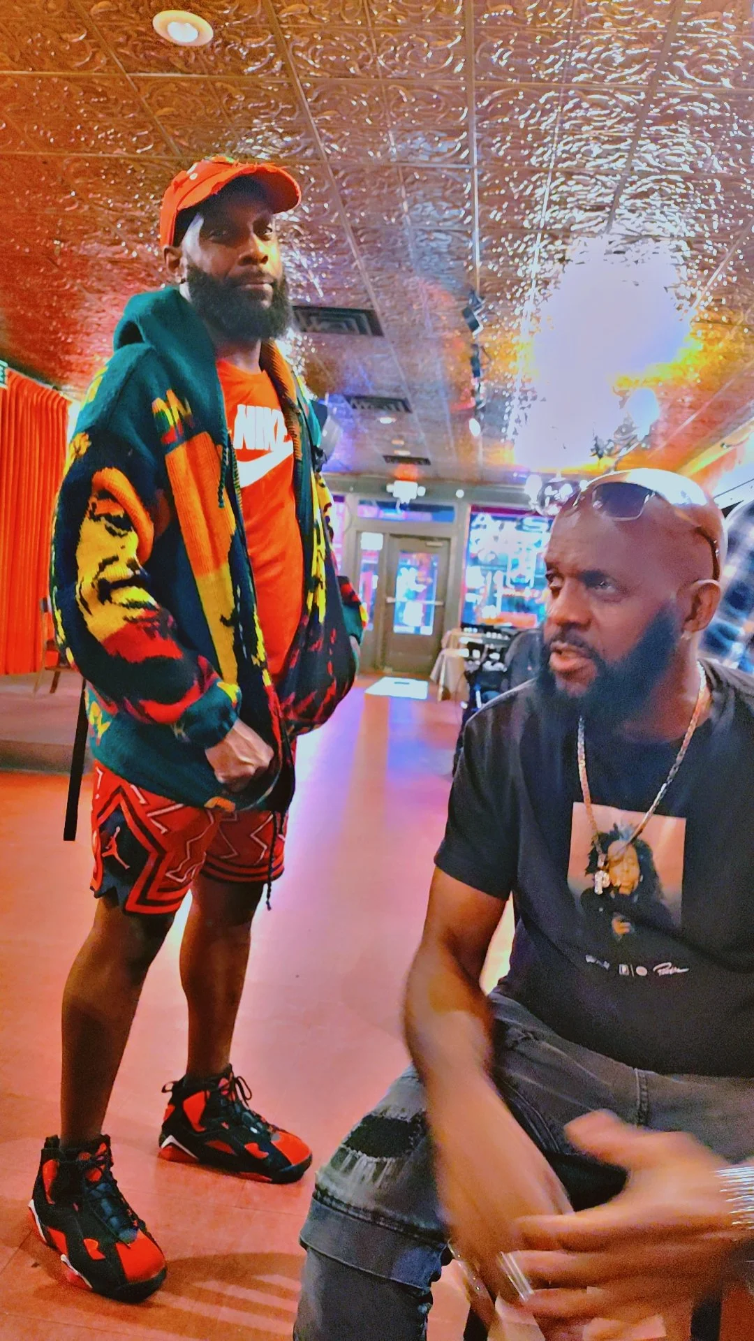 Two men in a dimly lit restaurant or bar, with one standing and wearing colorful streetwear, and the other sitting and wearing a black t-shirt, with neon lights and a decorated ceiling in the background.