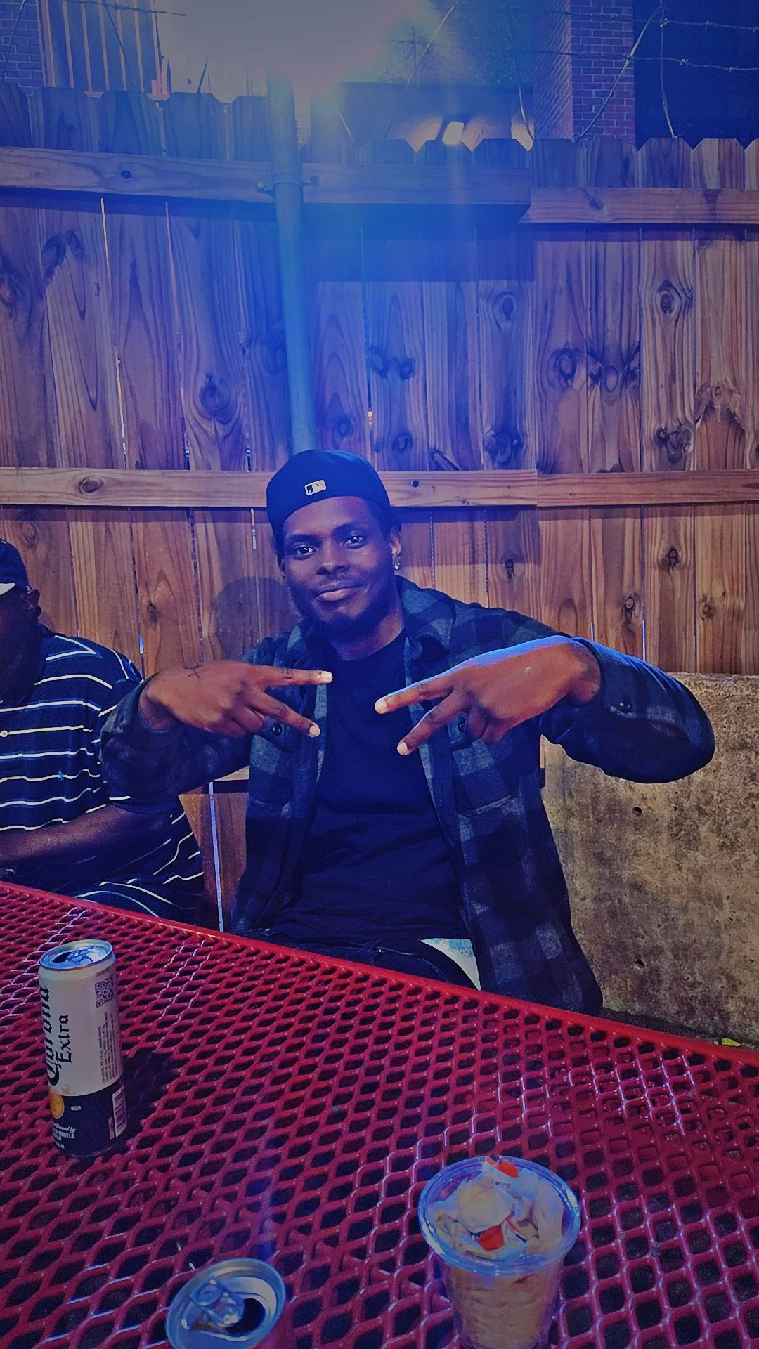 Young man wearing a black cap and plaid shirt making peace signs with both hands at an outdoor gathering, sitting at a red metal table with a soda can, snack container, and a drink.