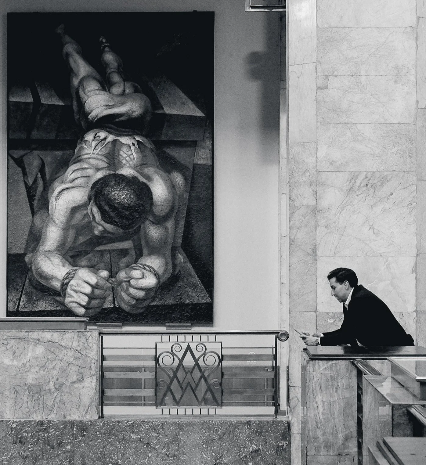 On the mezzanine level of the historic Palacio de Bellas Artes Museum in CDMC, in the foreground, a man leans against a marble wall with brass railing; his hands, eyes, and attention fixed on his phone. On the wall beside him hangs an unsettling im