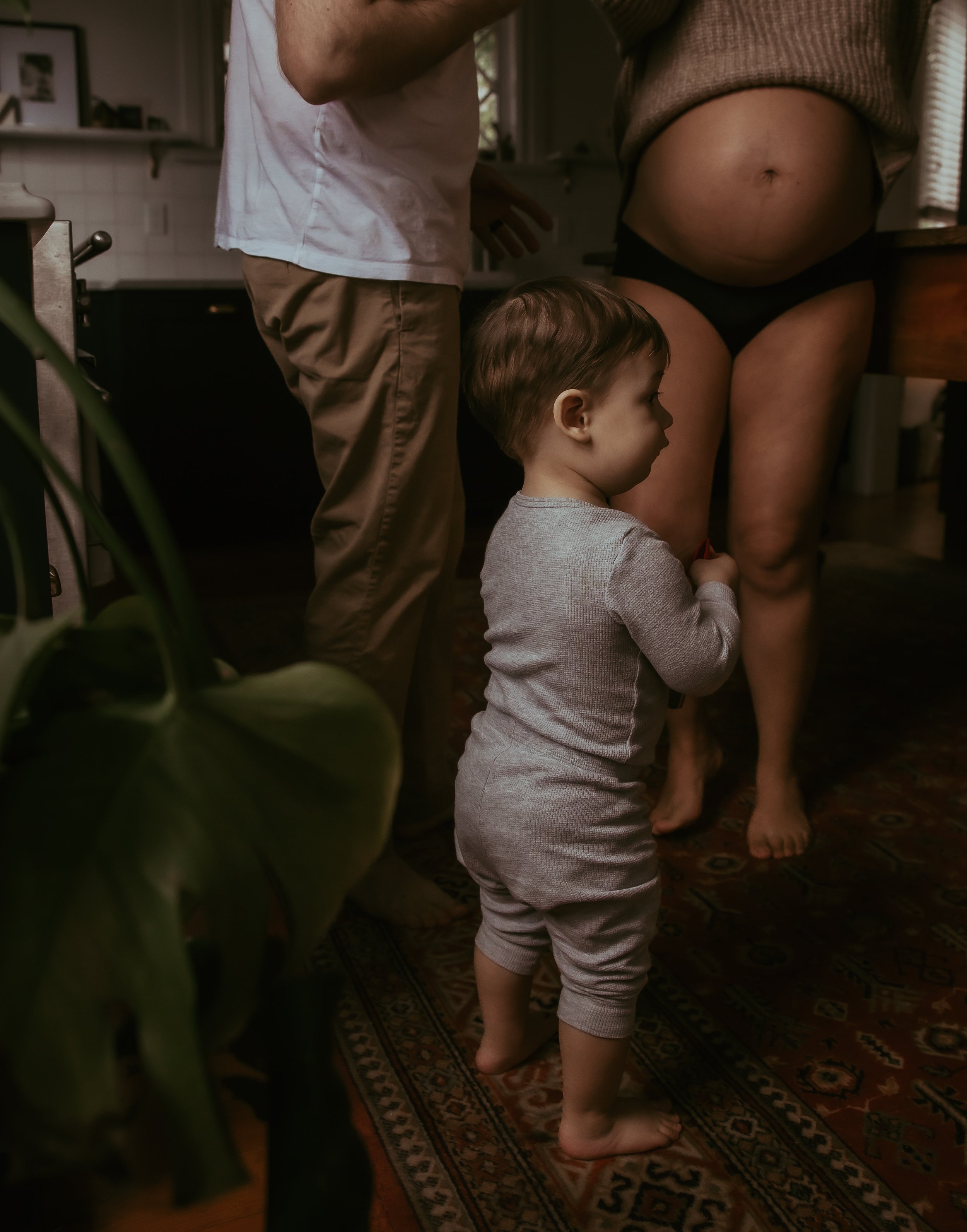 A young child in gray pajamas standing barefoot on a patterned rug, holding the leg of a pregnant woman with a visible belly, while another person stands nearby.