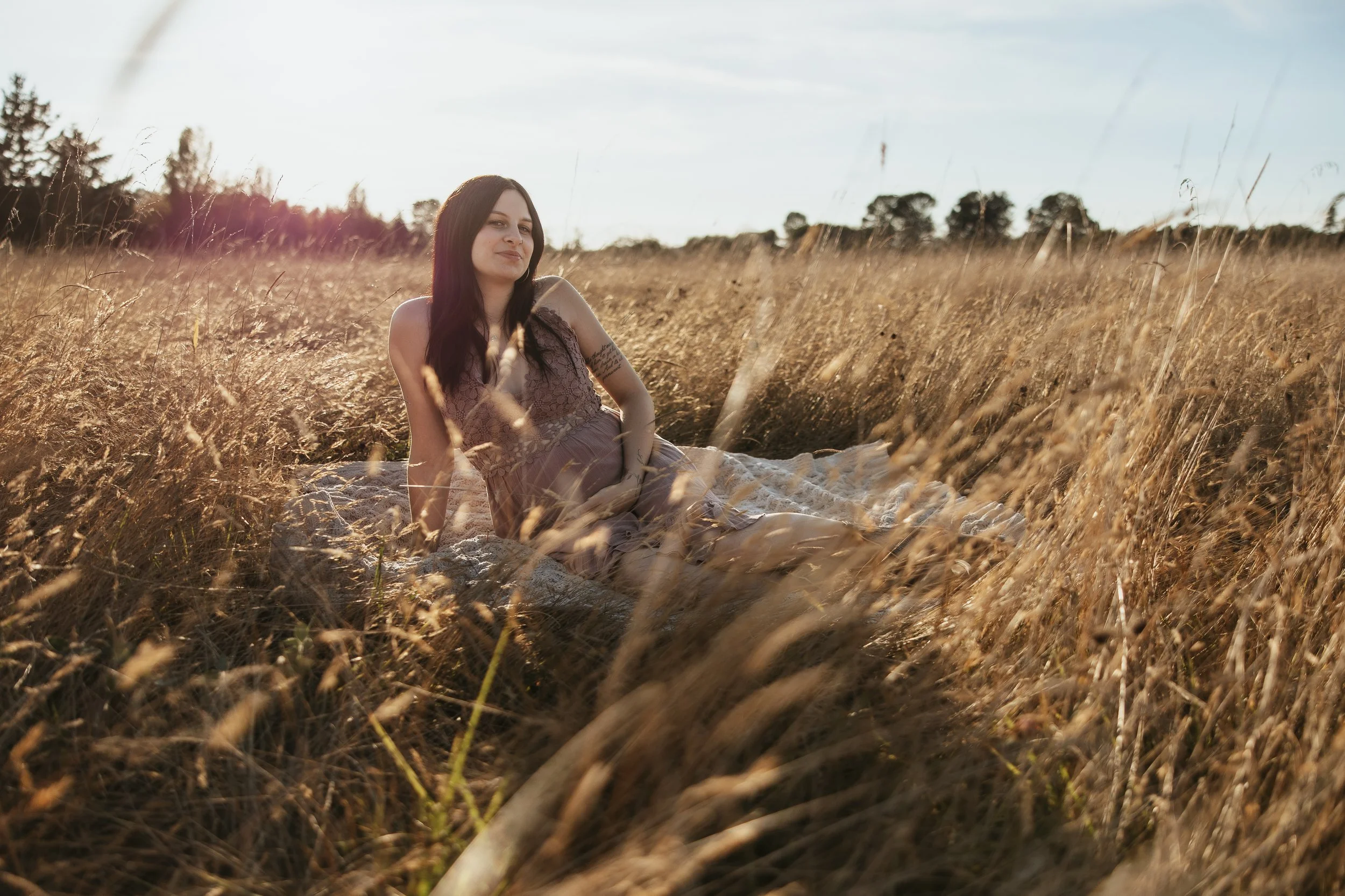 A pregnant mother sitting on a blanket in a field of tall, golden grass during sunset.