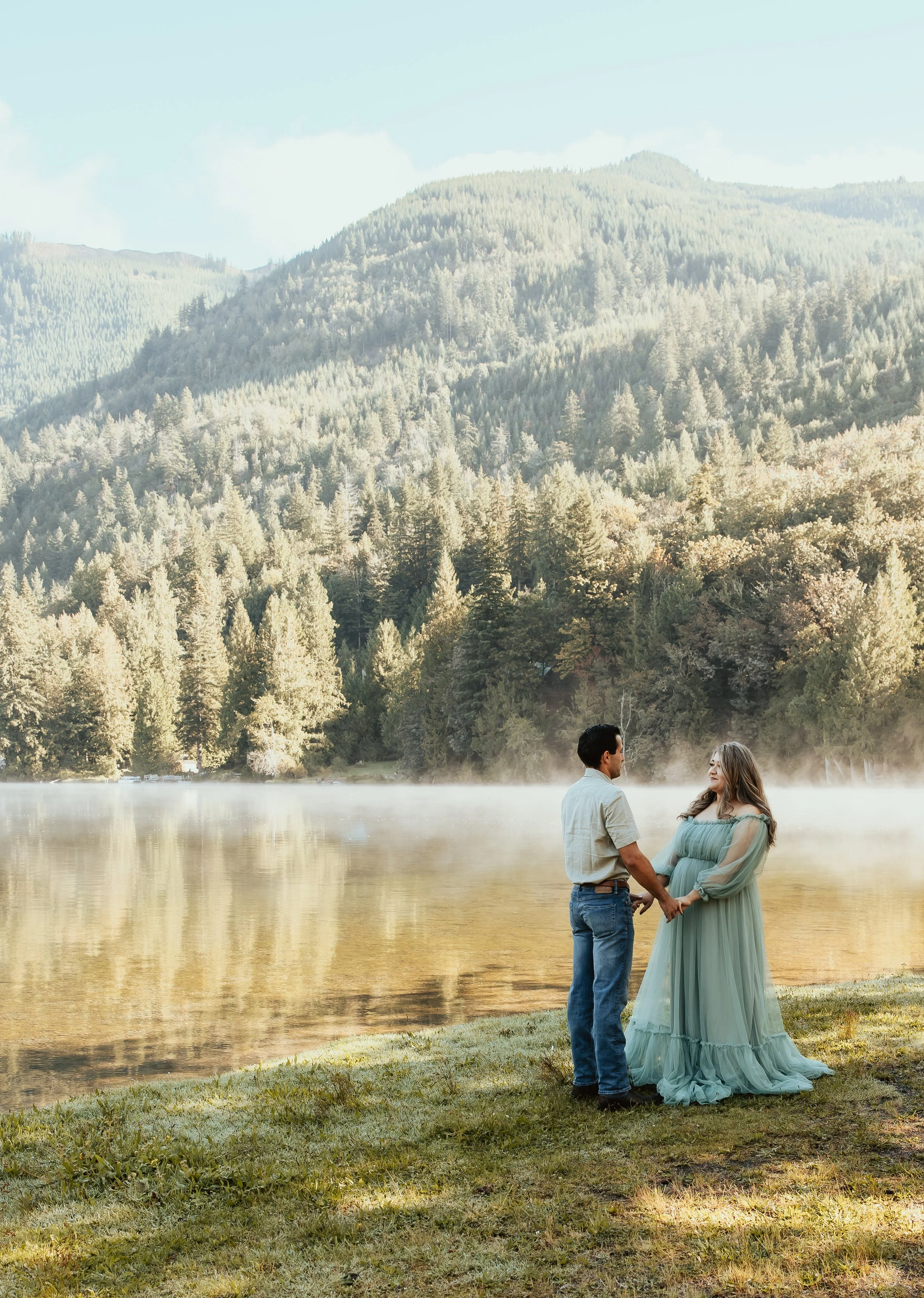 A couple holding hands by a lake in a forested mountainous area during daytime, with mist rising from the water.