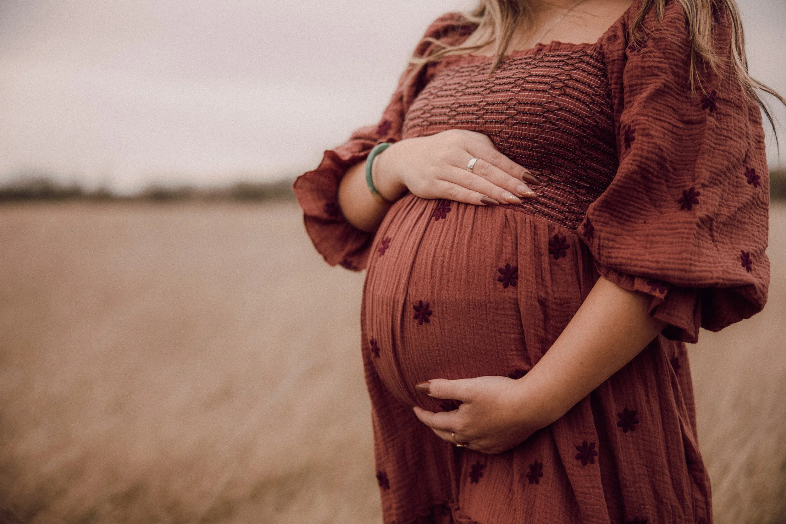 A pregnant woman wearing a rust-colored dress with floral embroidery, standing in a field, cradling her belly with both hands.