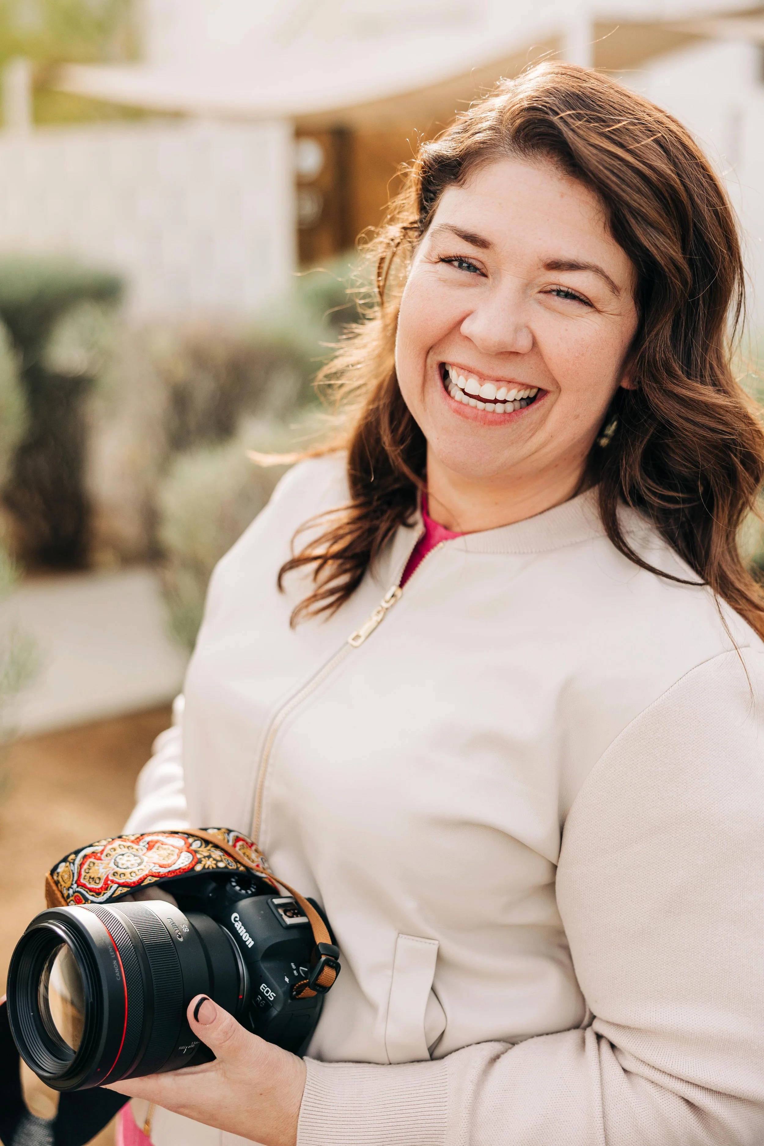A woman with medium-length brown hair smiling outdoors, holding a Canon DSLR camera.