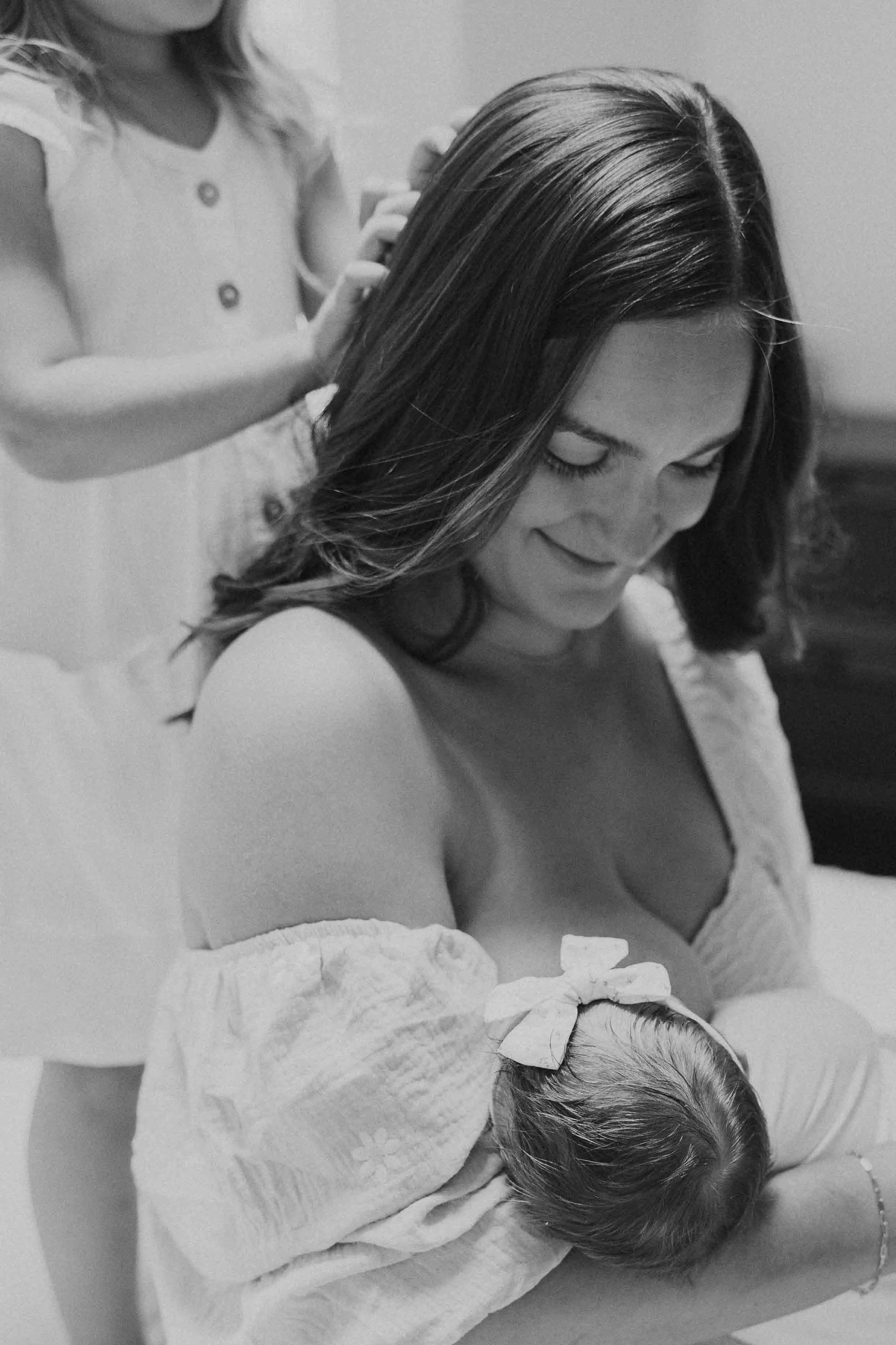 Mother breast feeding newborn while older daughter plays with her hair in in-home newborn photography session