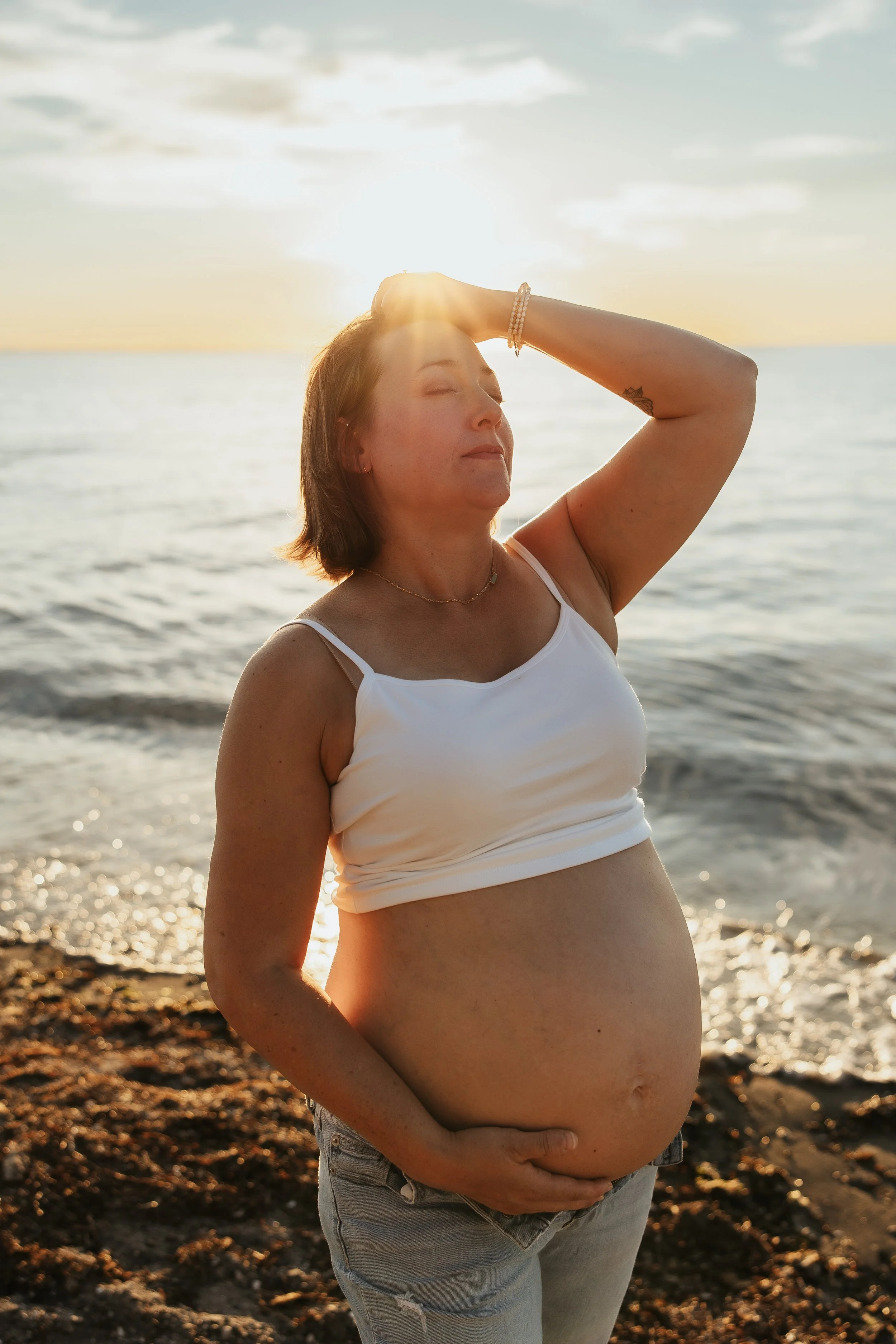 A pregnant woman stands on a beach at sunset, eyes closed, with her left hand on her belly and her right hand on her forehead, appearing peaceful and relaxed.