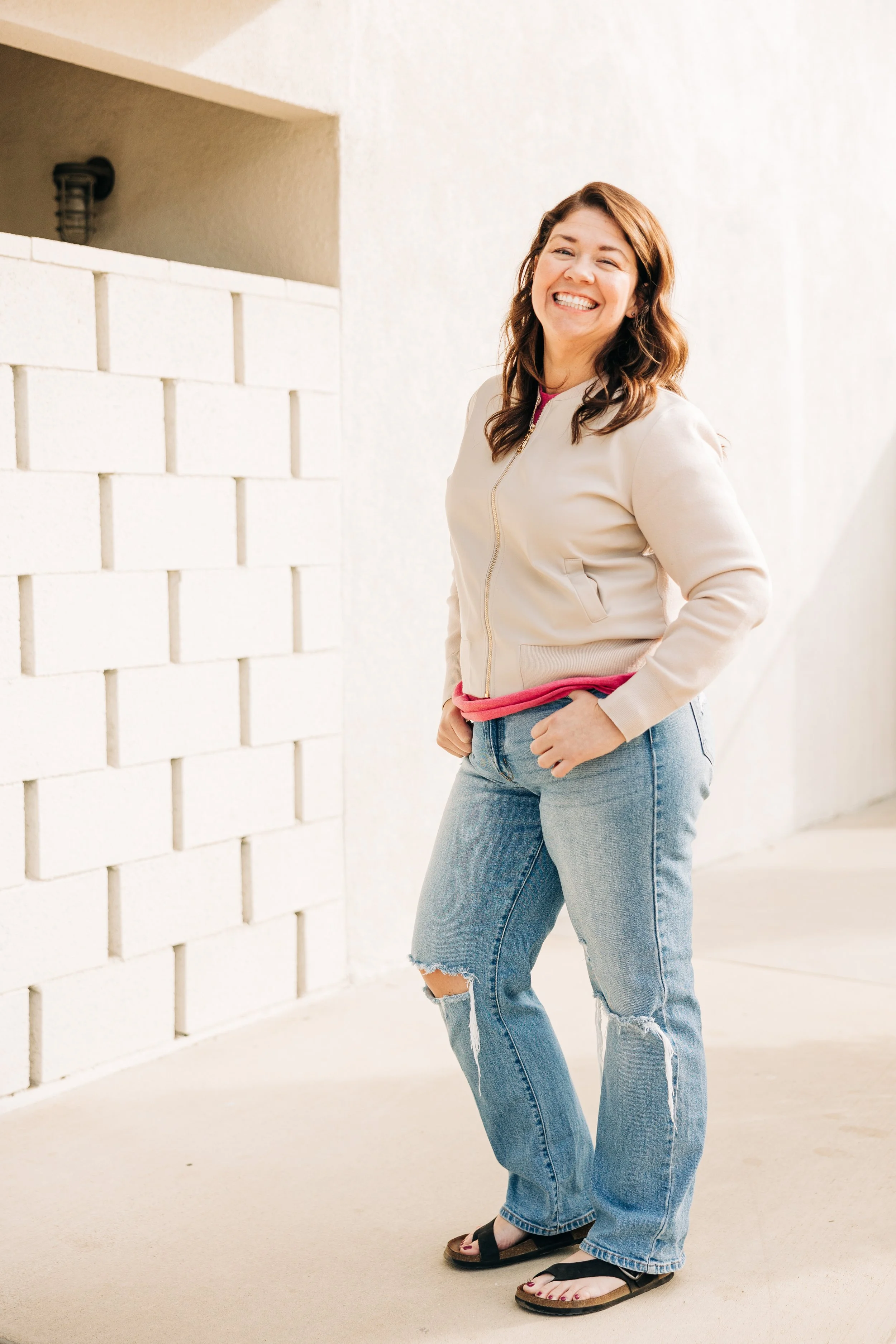 Smiling woman with brown hair, wearing a beige jacket, pink shirt, ripped jeans, and sandals, standing outdoors against a white wall.