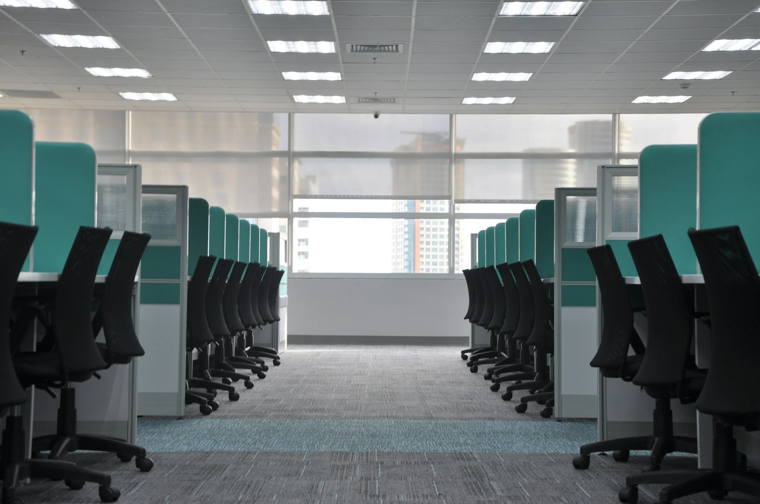Empty modern office space with rows of black office chairs and divided workstations with teal panels, large windows providing natural light, and a cityscape view in the background.
