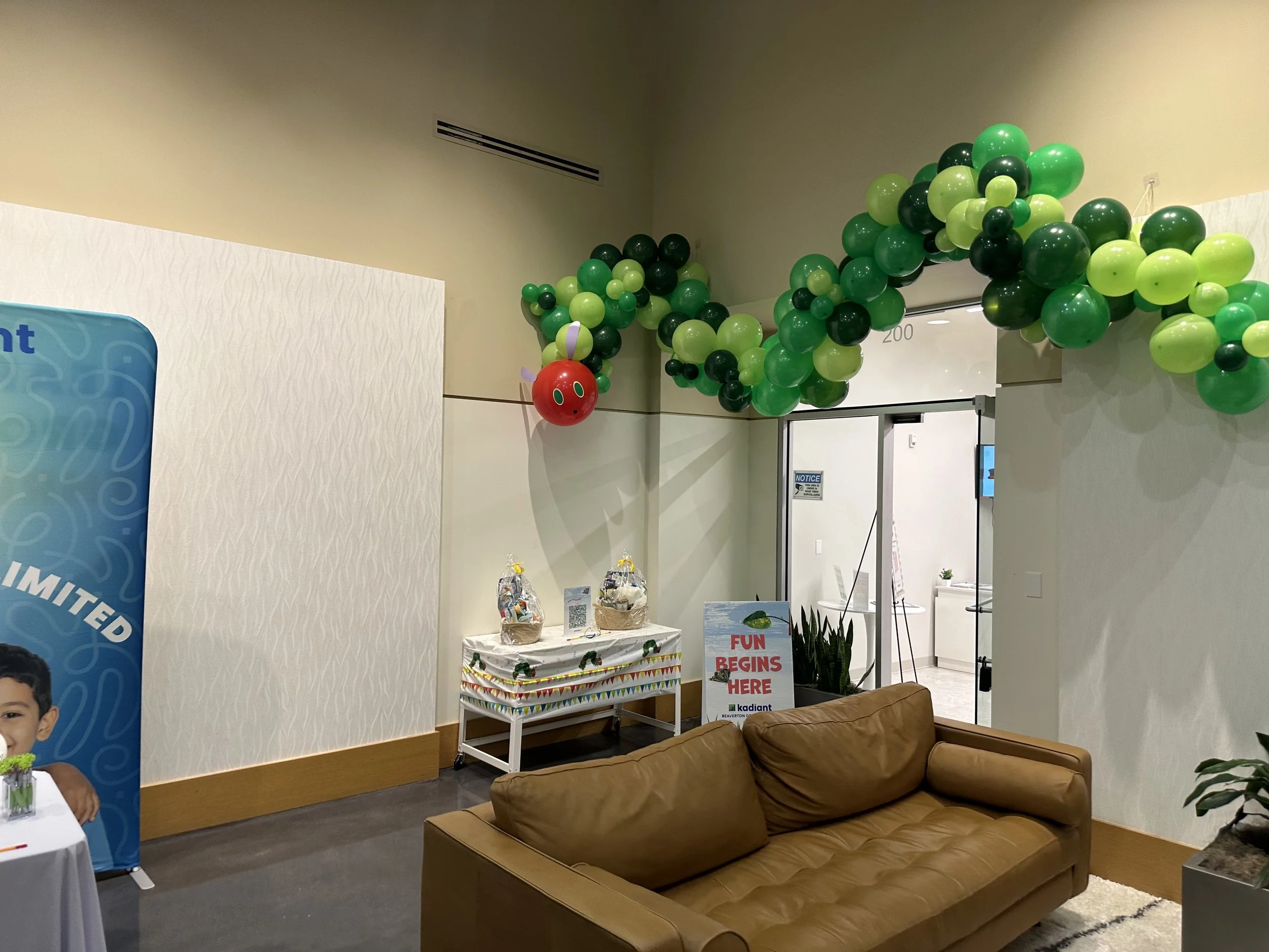 Indoor space decorated with green and black balloons forming an arch and a clown-shaped balloon on the left. A tan sofa is in the foreground, with a table holding gift baskets and a sign that reads 'Fun begins here'. Part of a poster with a child's image is visible on the left side.