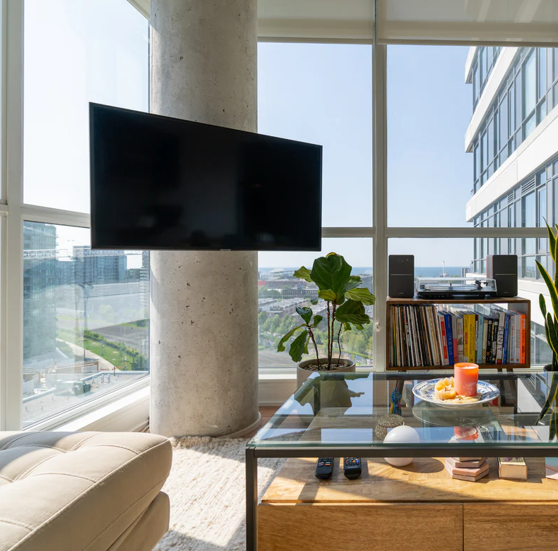 Modern living room with large windows, flat-screen TV, potted plant, bookshelf with vinyl records, glass-top table, decorative items, and city view.
