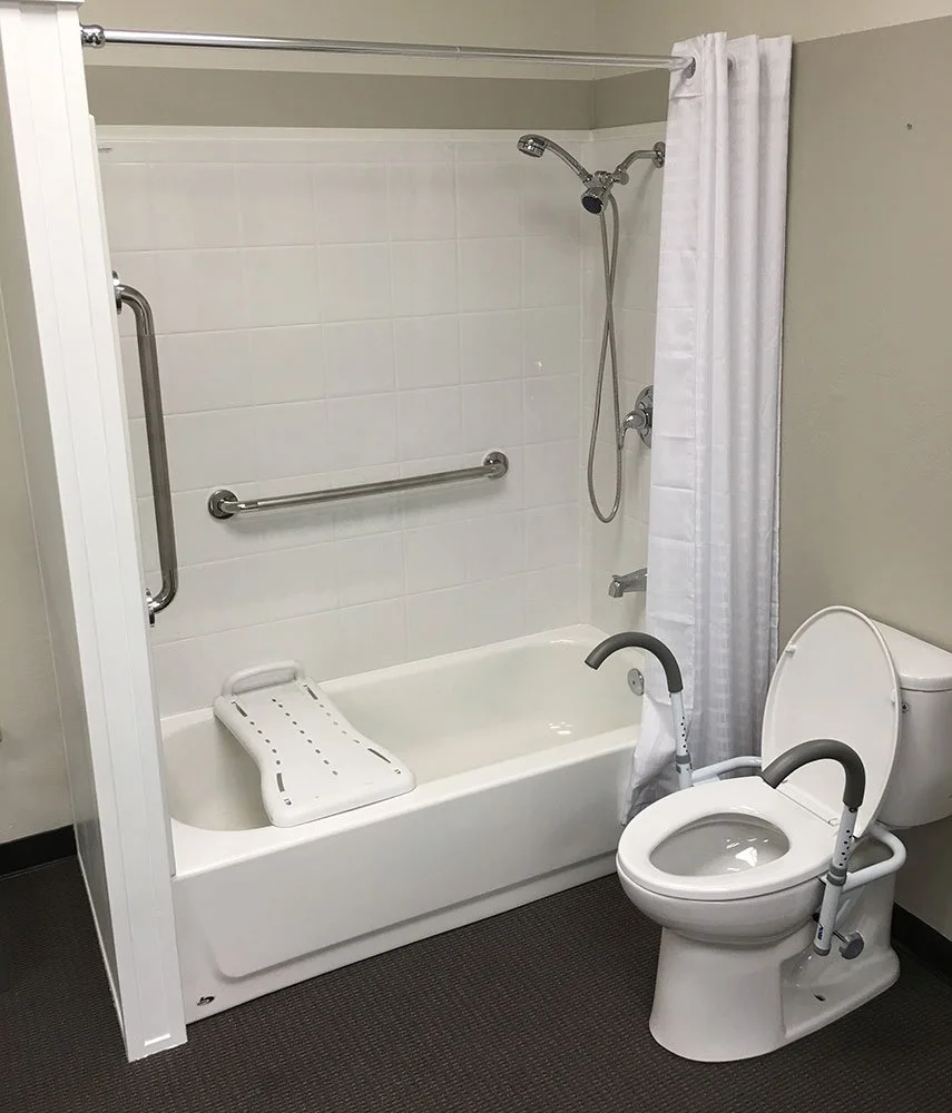 Accessible bathroom with a bathtub, grab bars, showerhead, toilet with safety rails, and a shower curtain.