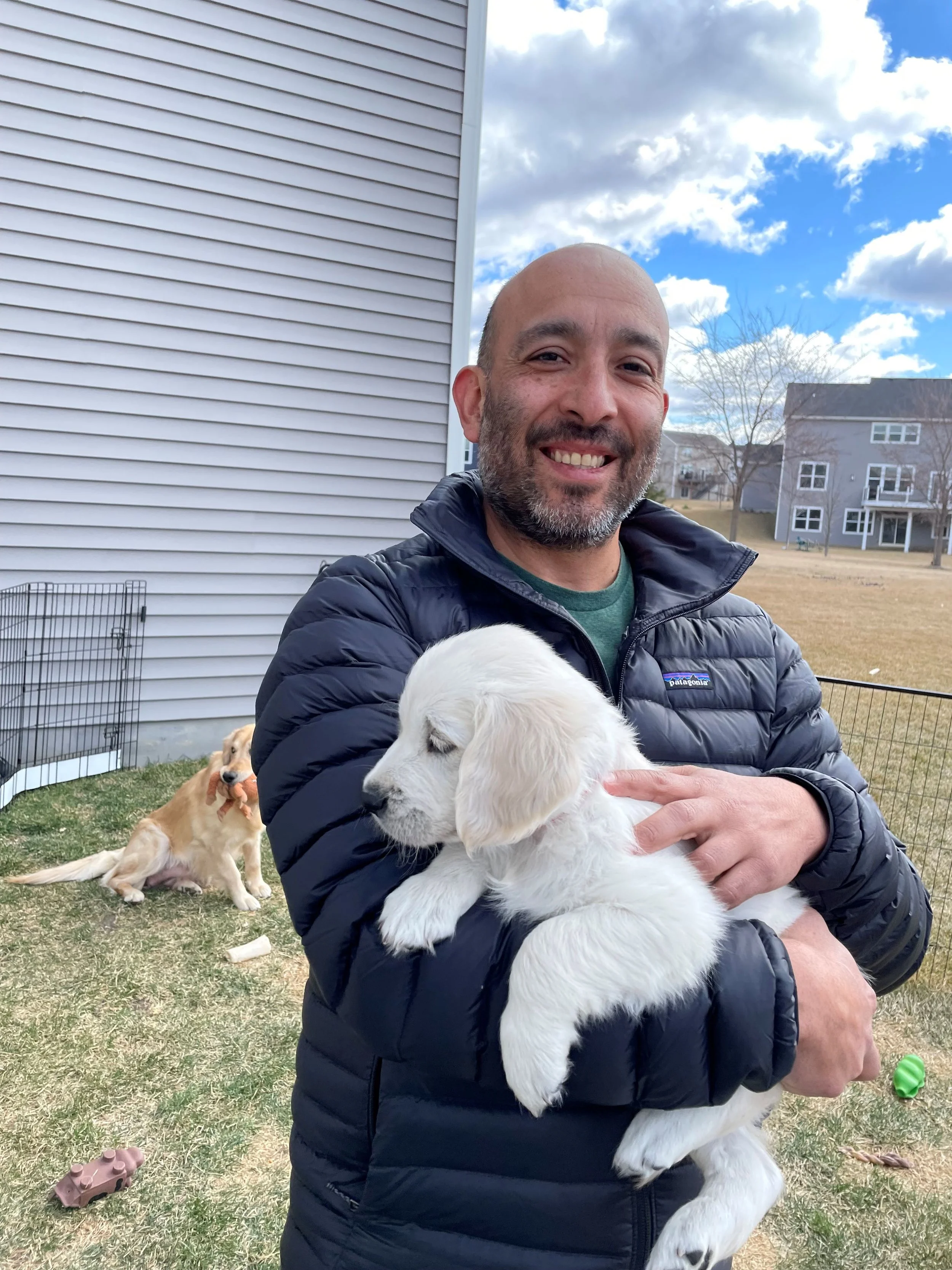 A man holding a white puppy outdoors with a house and another dog in the background.