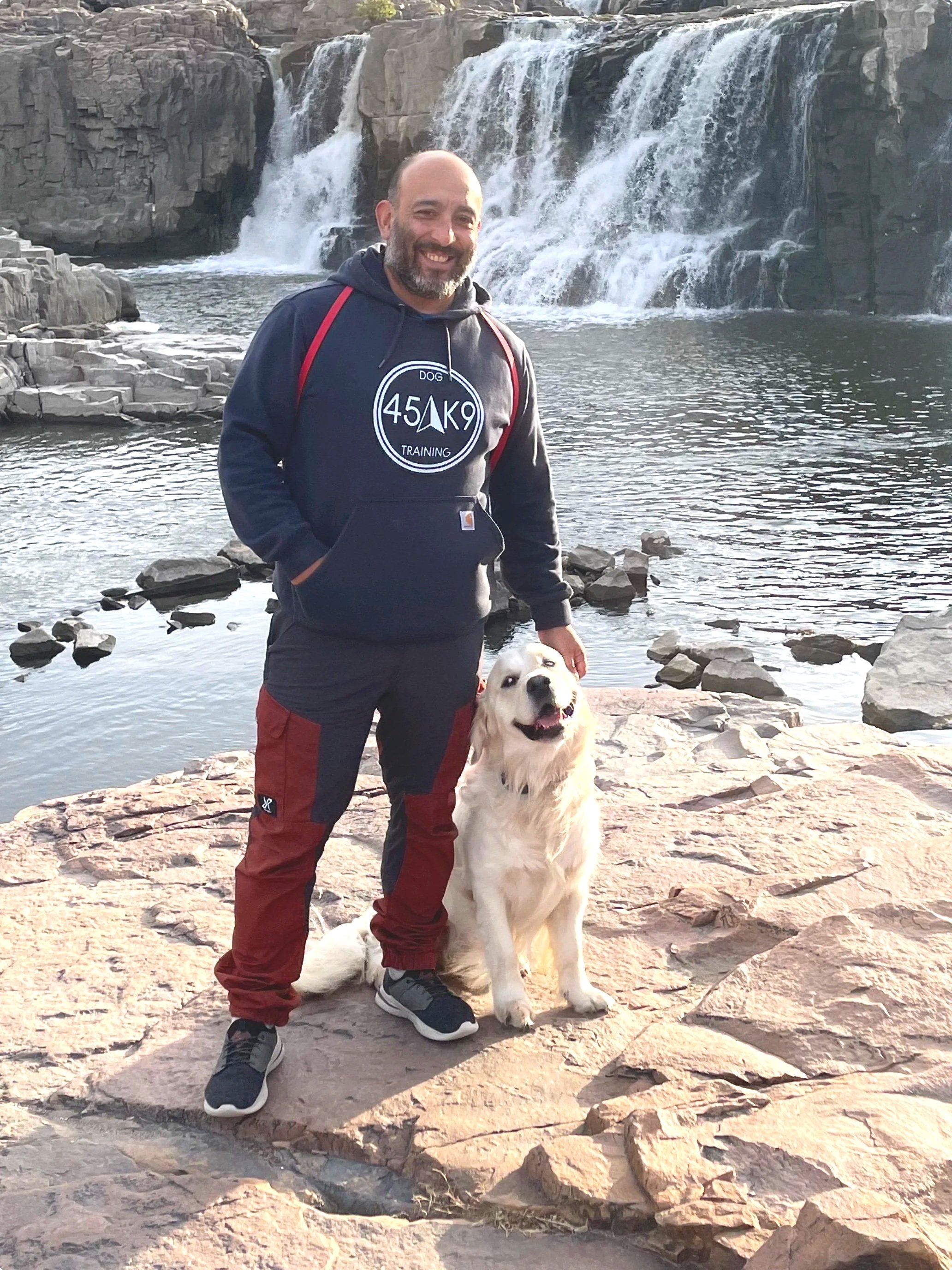 A man and a golden retriever dog standing by a waterfall, on a rocky shoreline. The man is smiling, wearing a navy hoodie with a logo, red and gray pants, and sneakers. The dog is sitting, looking happy, with its tongue out.