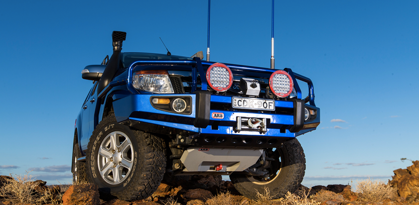 Blue off-road vehicle on rocky terrain with additional front lights and a custom bumper, under a clear blue sky.