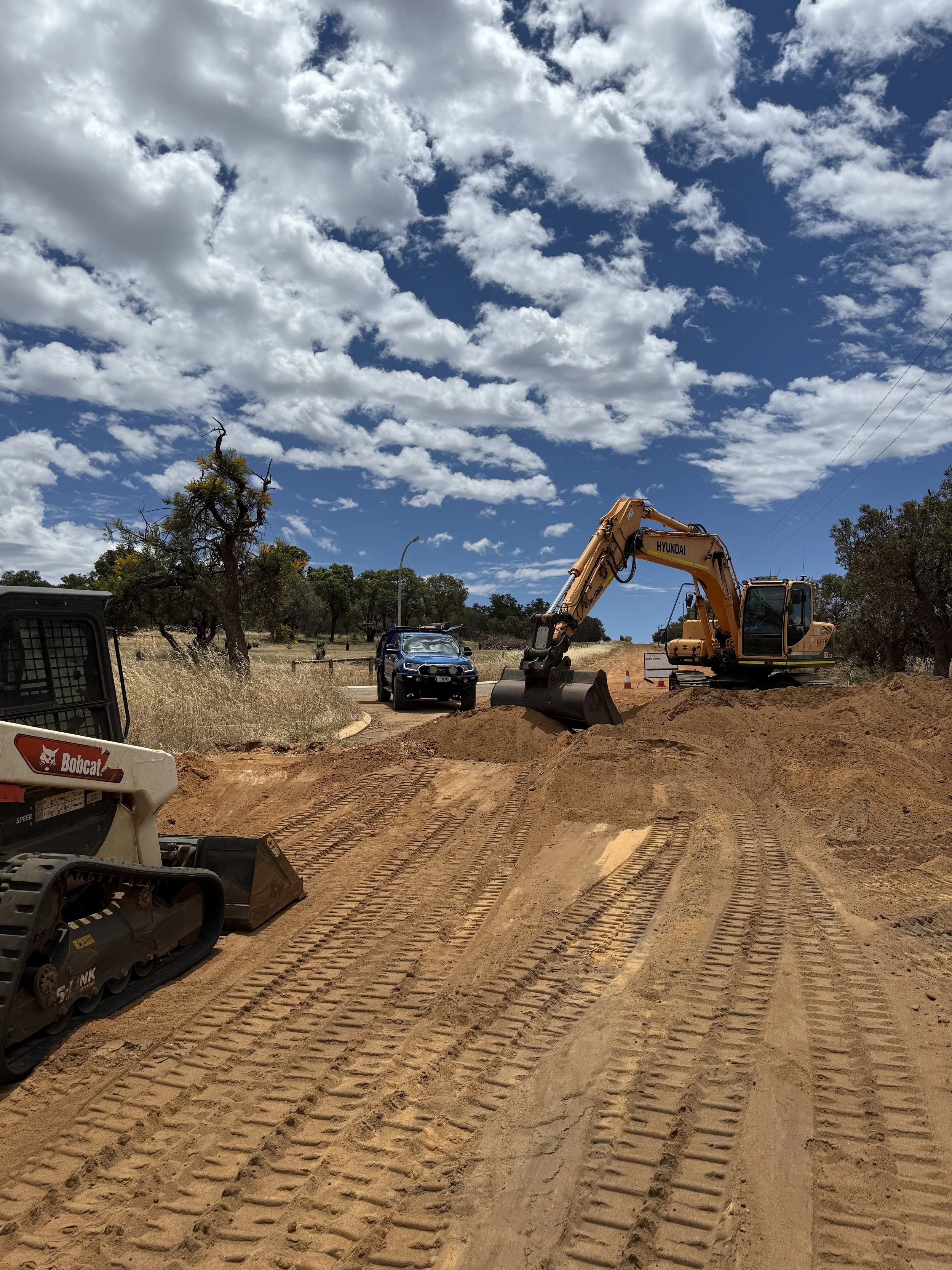 Construction site with a bulldozer and a tracked vehicle on dirt with tire tracks, a black car in the background, trees, and a partly cloudy sky.