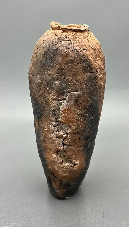 An ancient, weathered clay or ceramic vessel with an irregular shape and a rough surface, featuring a face-like indentation on its front.